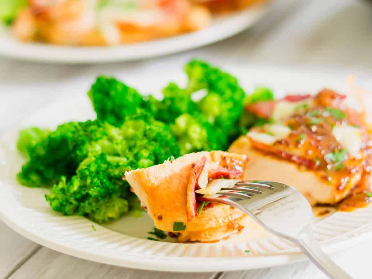 A white plate with baked chicken topped with sauce and herbs, accompanied by steamed broccoli. A fork holds a slice of chicken, revealing a bit of bacon and cheddar inside. Another similar plate is blurred in the background.