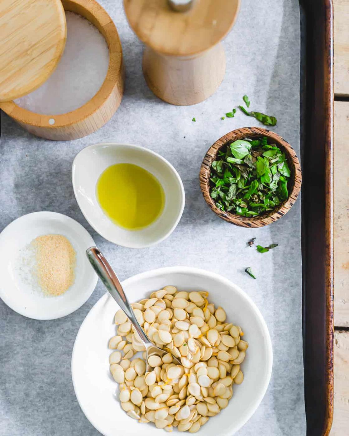 Delicata squash seeds in a bowl with ingredients to roast: extra virgin olive oil, garlic powder, fresh herbs, salt and pepper.