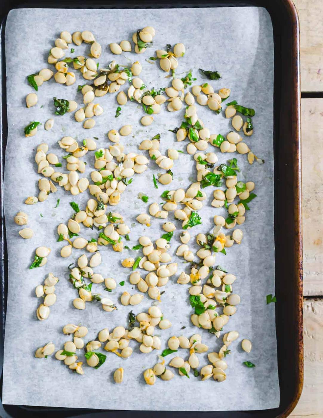 Delicata squash seeds with olive oil, garlic powder, fresh minced herbs, salt and pepper spread out on a baking sheet.