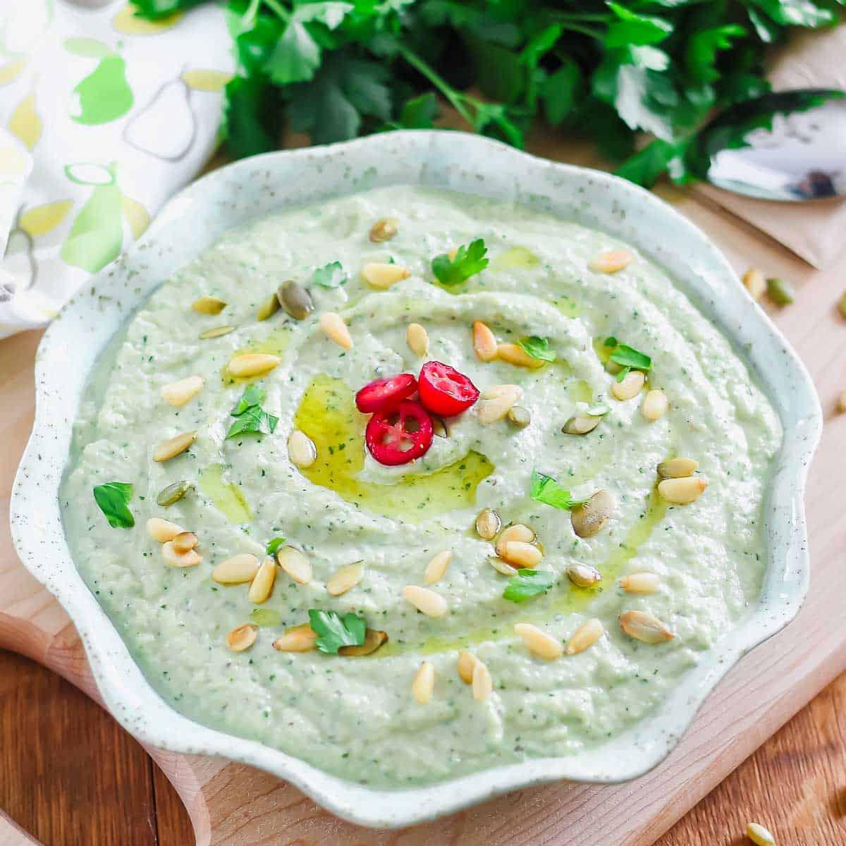 A bowl of roasted eggplant dip garnished with pine nuts, pumpkin seeds, and parsley. Three slices of red chili pepper are at the center. Fresh parsley and a patterned cloth are in the background.