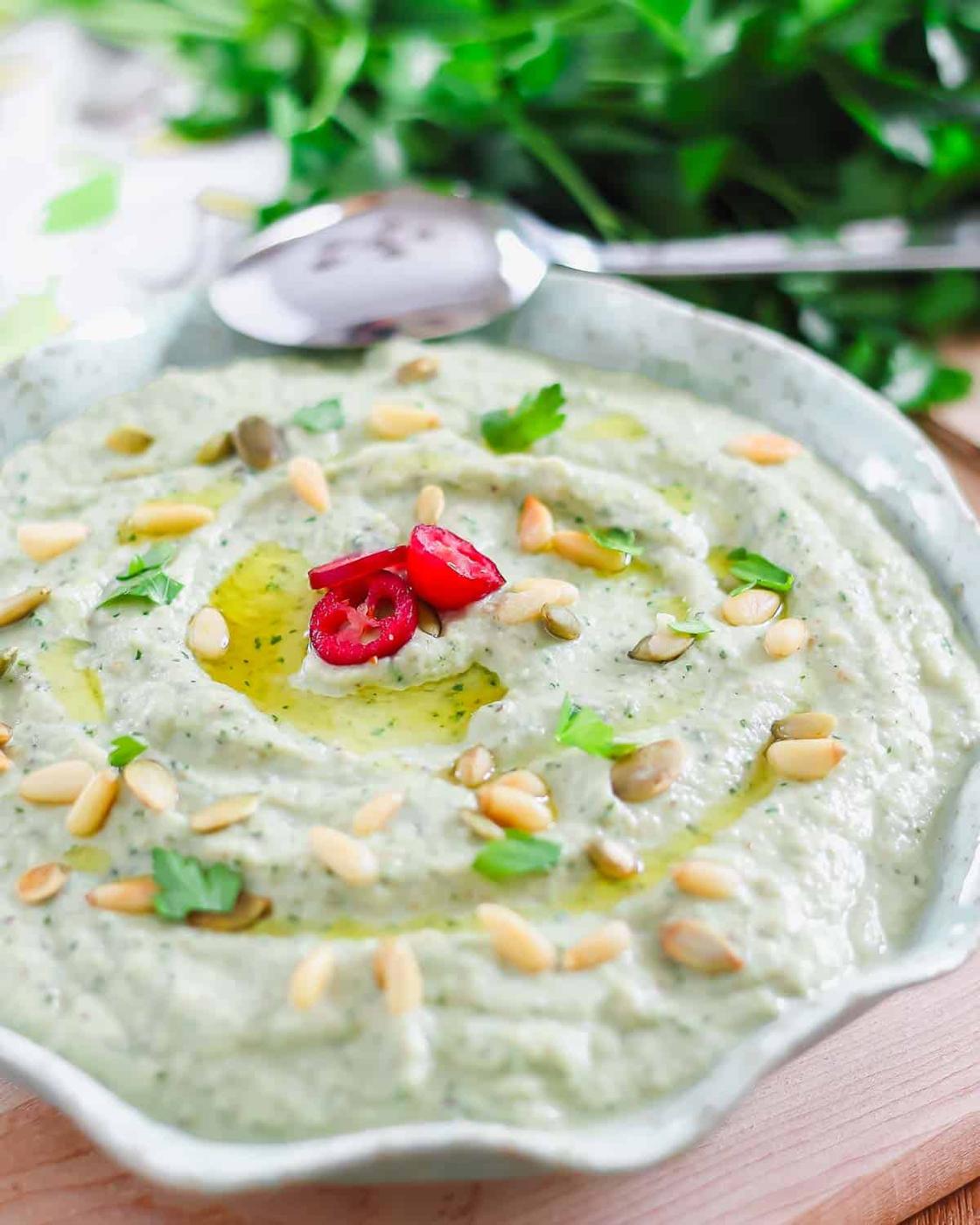A bowl of eggplant dip garnished with olive oil, sliced red chili, pine nuts, pumpkin seeds, and fresh parsley. A spoon rests beside it, with more fresh parsley in the background.