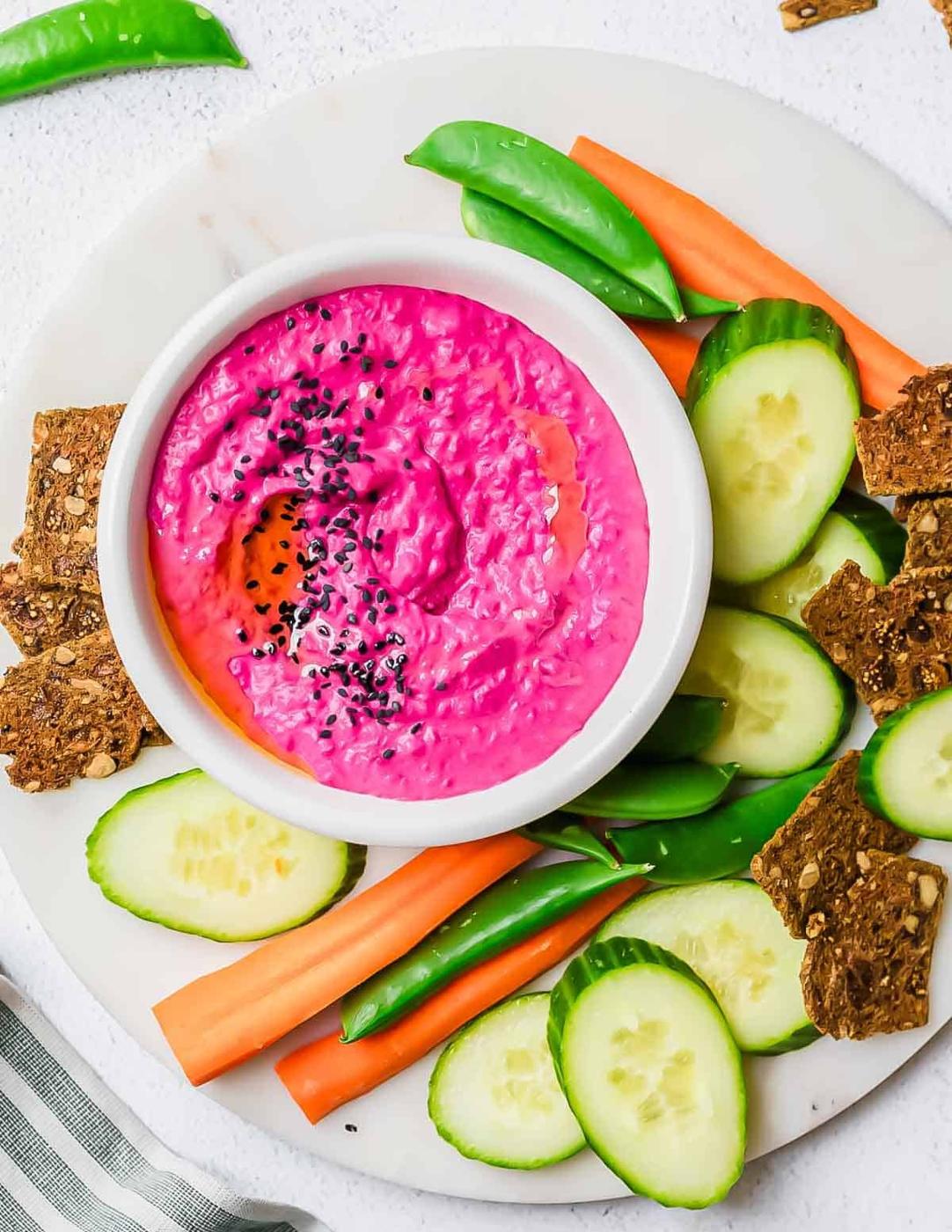 A bowl of bright pink beet hummus topped with black sesame seeds, surrounded by cucumber slices, carrot sticks, snap peas, and seeded crackers on a white round platter.