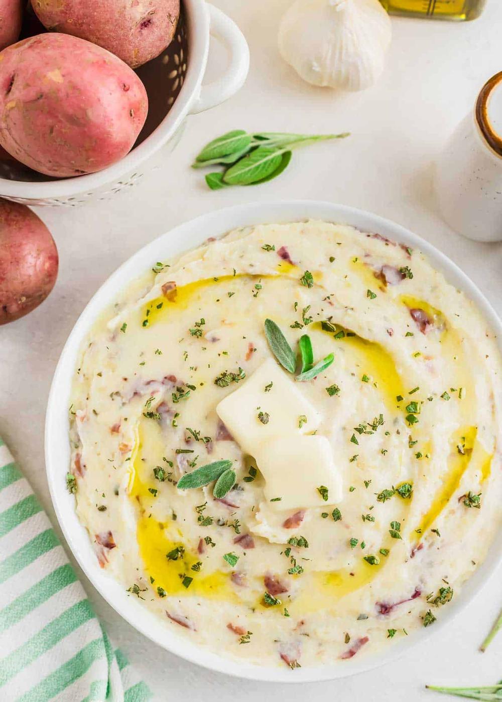A bowl of roasted garlic mashed potatoes topped with melting butter and garnished with fresh herbs. Red potatoes and a garlic bulb are on the side. A green-striped cloth and a small dish of salt are nearby.