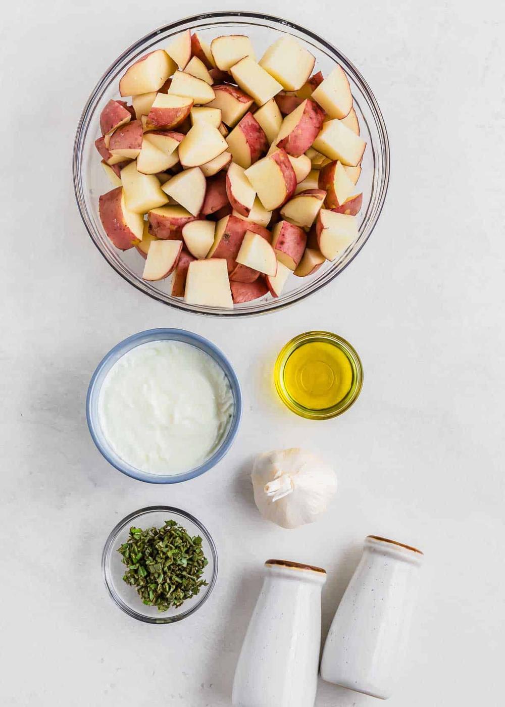 A glass bowl filled with chopped red potatoes, a small bowl of yogurt, a small bowl of chopped herbs, a bulb of garlic, a small bowl of olive oil, and two salt and pepper shakers are arranged on a light countertop.