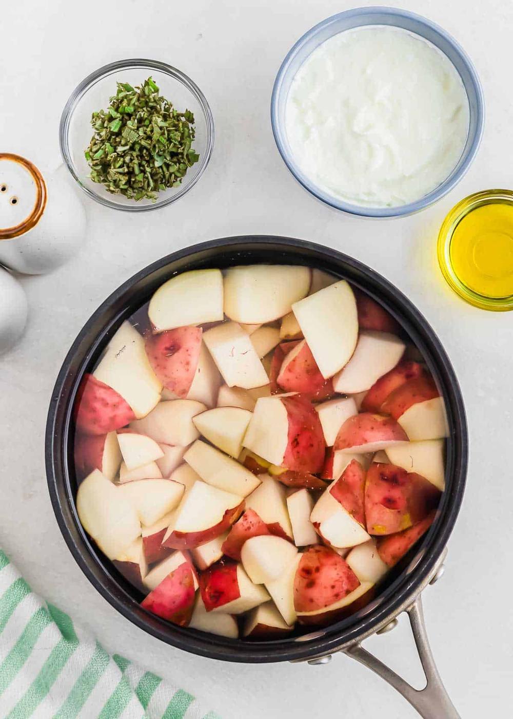 A pot filled with chopped red potatoes submerged in water. Surrounding the pot are small bowls containing sour cream, chopped herbs, olive oil, and a set of salt and pepper shakers on a white surface. A striped green cloth is nearby.