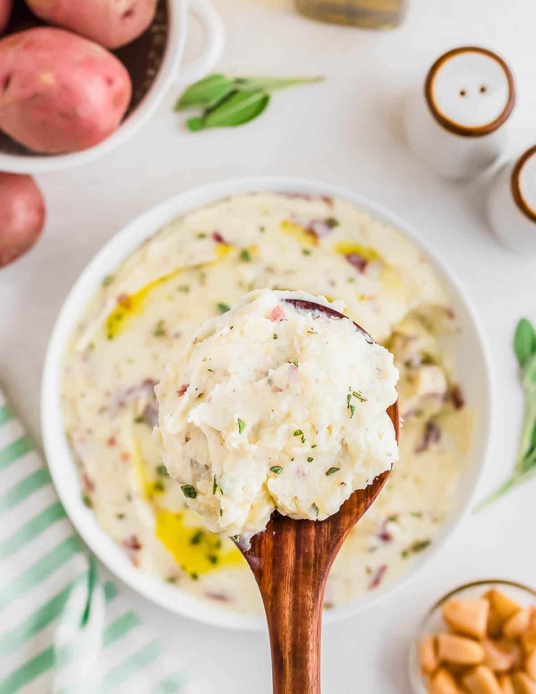 A wooden spoon holds a scoop of creamy mashed potatoes garnished with herbs and roasted garlic. Below, a bowl of mashed potatoes is drizzled with oil. Nearby are red potatoes, salt and pepper shakers, and a striped cloth.