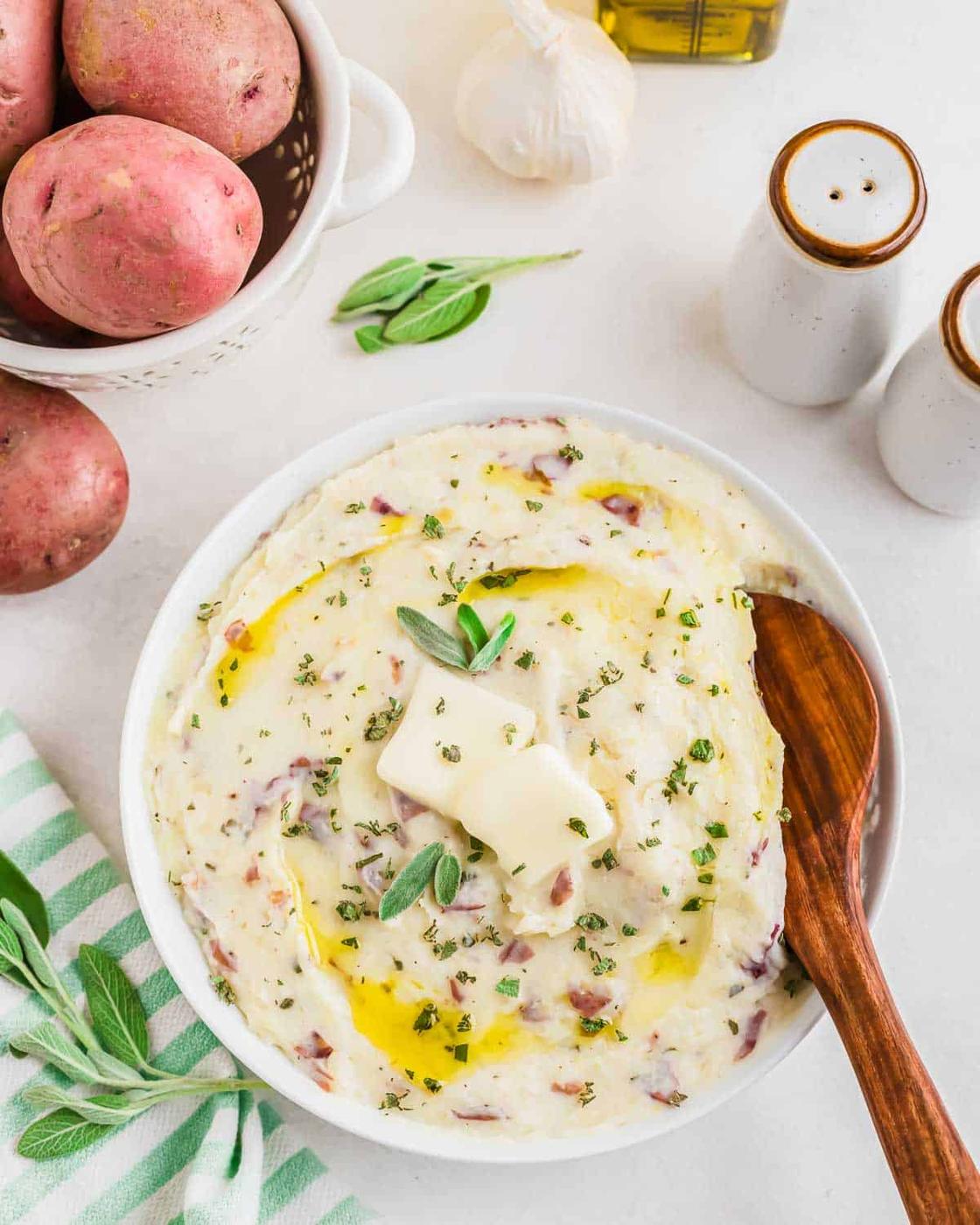 A bowl of creamy roasted garlic mashed potatoes with skins, garnished with herbs and two pats of butter, sits on a table. A wooden spoon is placed inside the bowl. Nearby are whole red potatoes, garlic, salt and pepper shakers, and a striped cloth.
