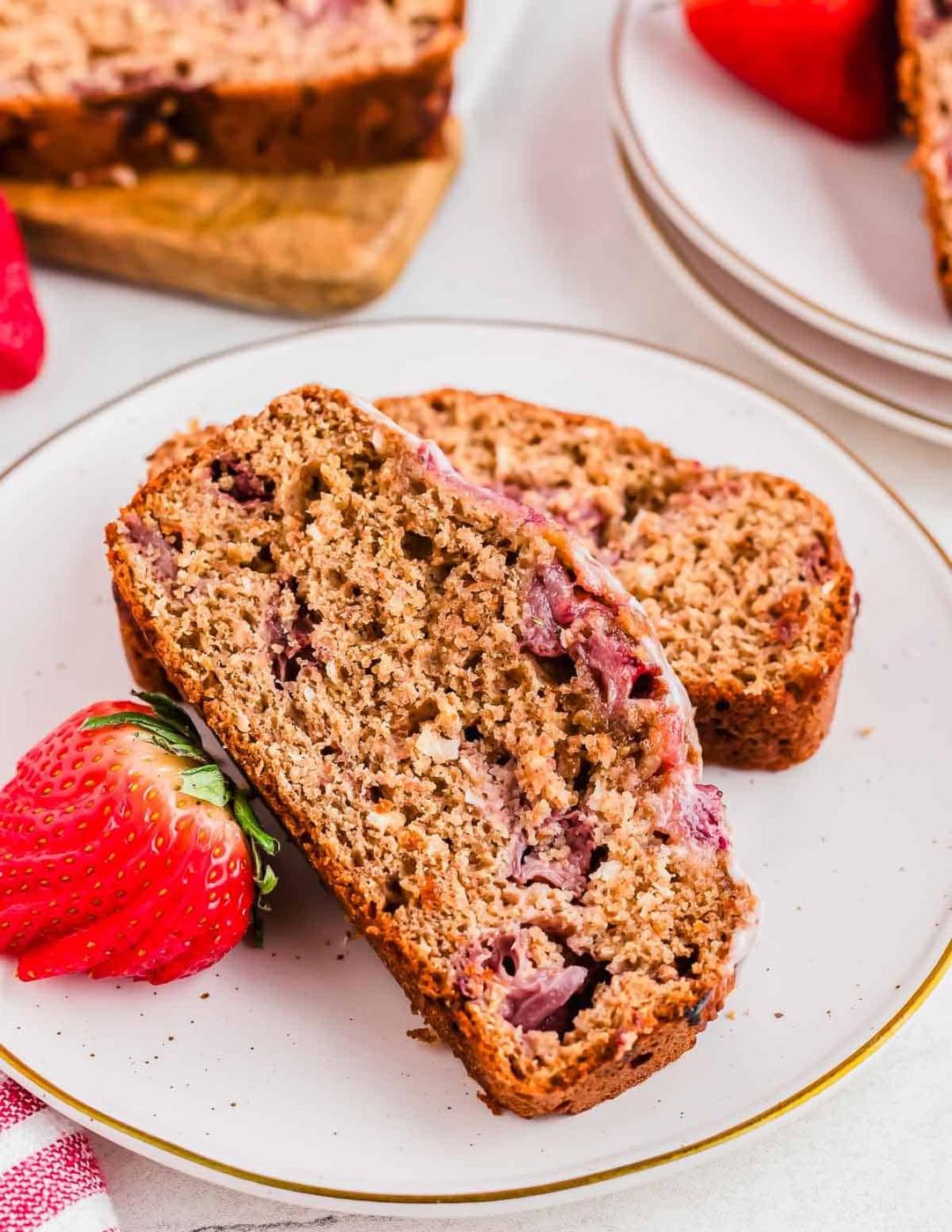 Two slices of strawberry bread are placed on a white plate, accompanied by a fresh strawberry. The bread features visible chunks of strawberries and a moist, dense texture. Another plate and a cutting board with more bread are in the background.