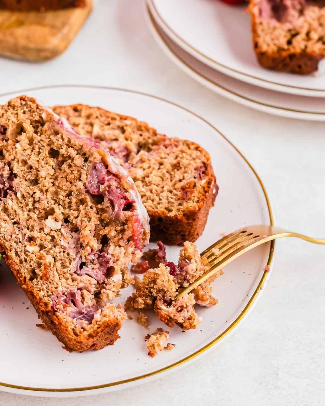 Slices of a crumbly moist strawberry bread with visible pieces of fruit, are placed on a white plate with a gold trim. A fork rests on the plate, breaking off a piece of the bread. The background includes another plate and blurred elements.