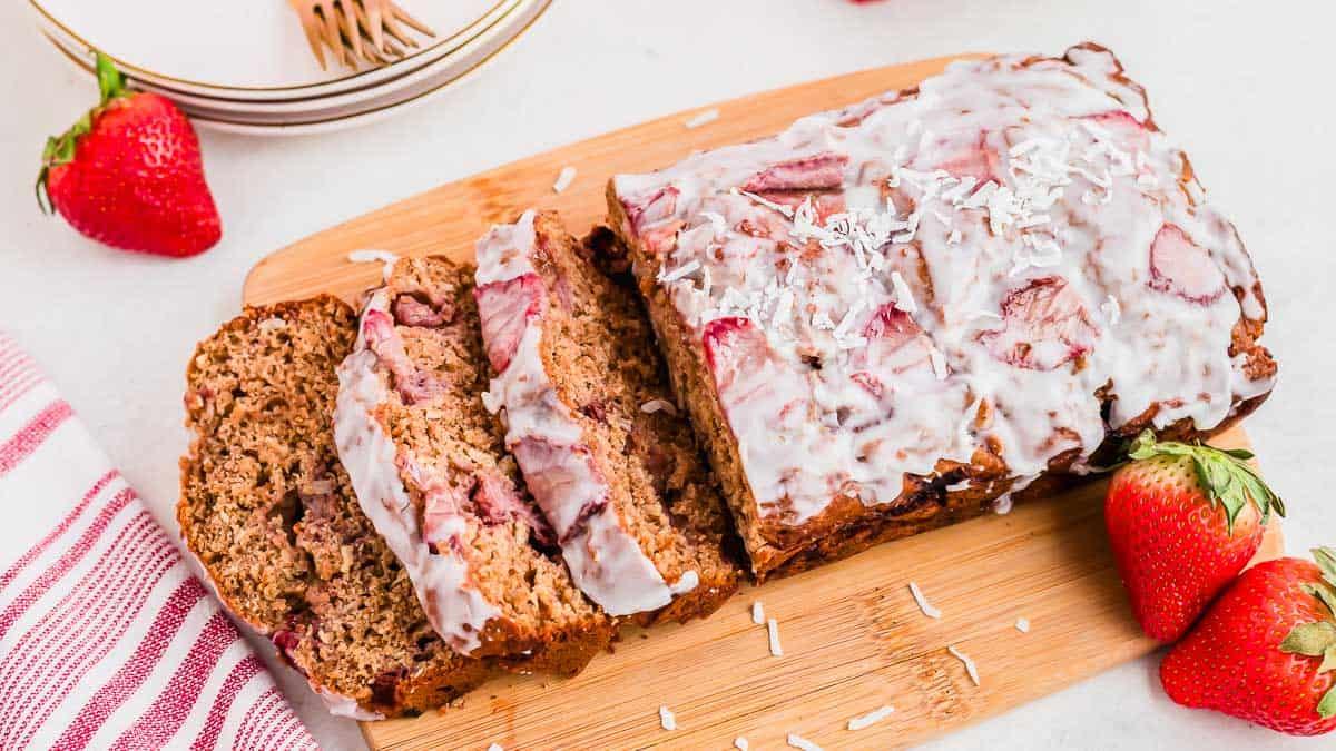 A sliced loaf of strawberry bread with icing on a wooden cutting board. Surrounded by fresh strawberries and a red-striped cloth. Some shredded coconut sprinkled on top adds texture to the glaze. White plates and gold forks are in the background.