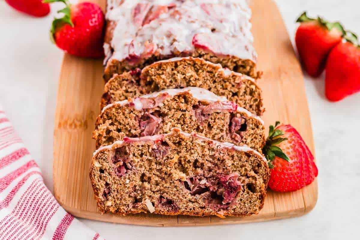 Sliced strawberry bread with glaze on a wooden cutting board, garnished with fresh strawberries. A red and white striped cloth napkin is visible on the side.