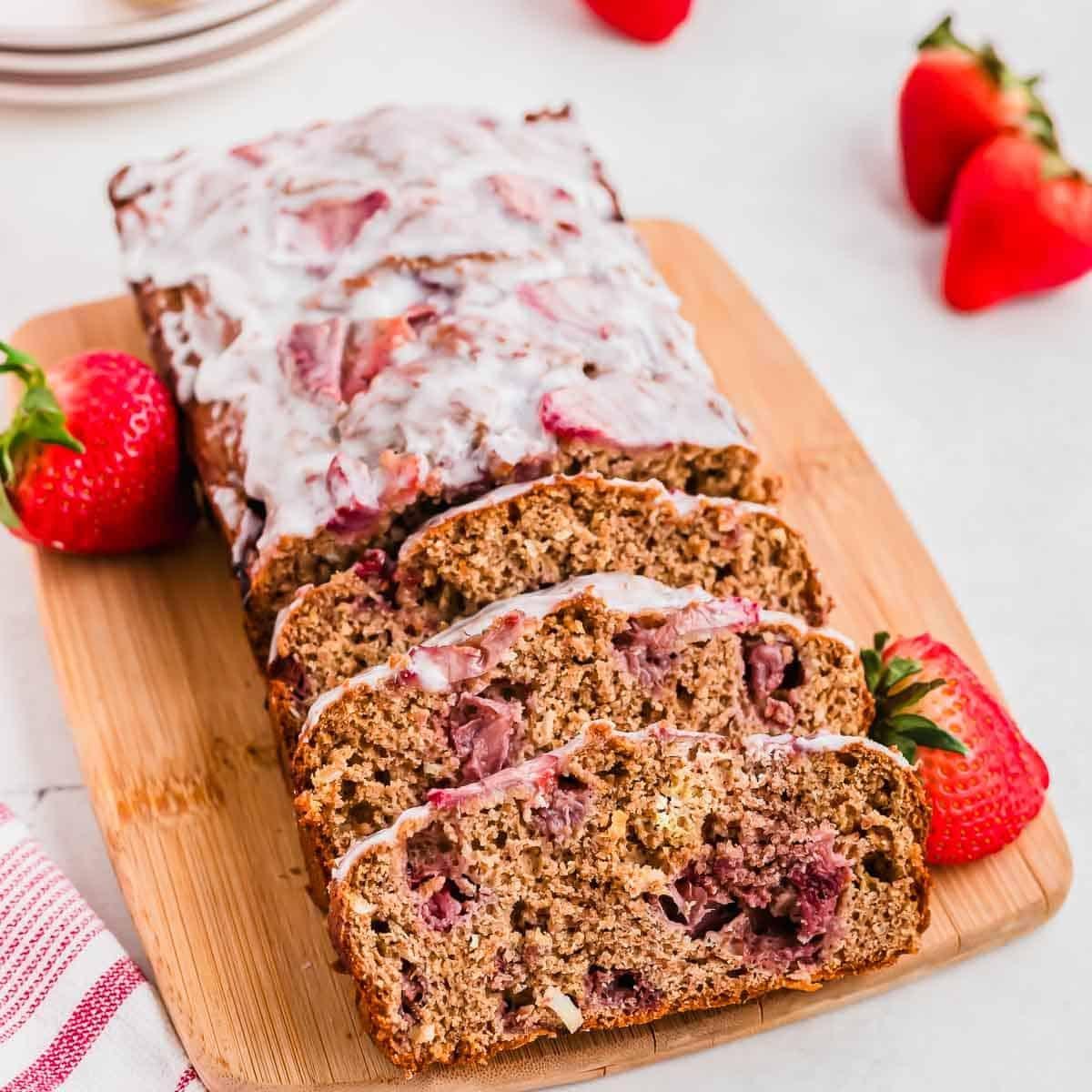 A sliced loaf of strawberry bread with glaze on a wooden board, surrounded by whole strawberries. A red and white striped cloth is in the foreground, and a stack of plates is partially visible in the background.