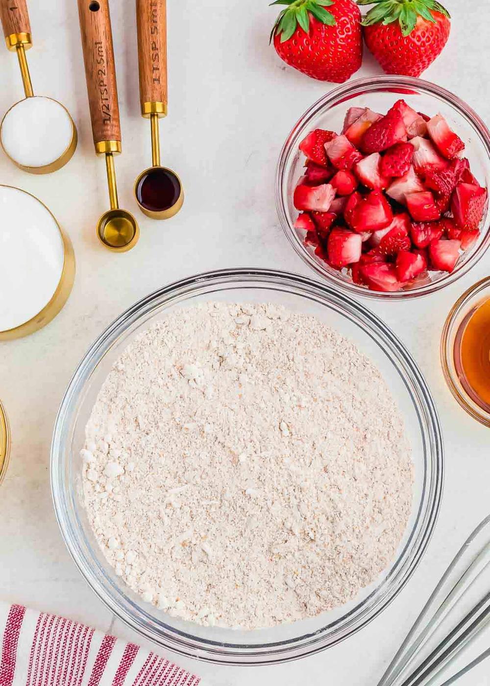 Top view of baking ingredients, including a bowl of flour, a bowl of chopped strawberries, measuring spoons, a cup of sugar, a cup of brown liquid, and whole strawberries. A striped cloth is partially visible at the bottom.