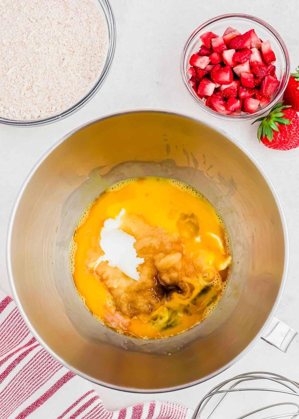 A mixing bowl contains eggs, applesauce, vanilla and other wet ingredients. Surrounding the bowl are ingredients: a bowl of diced strawberries, another with flour, and a red-striped kitchen towel, suggesting preparation for baking.