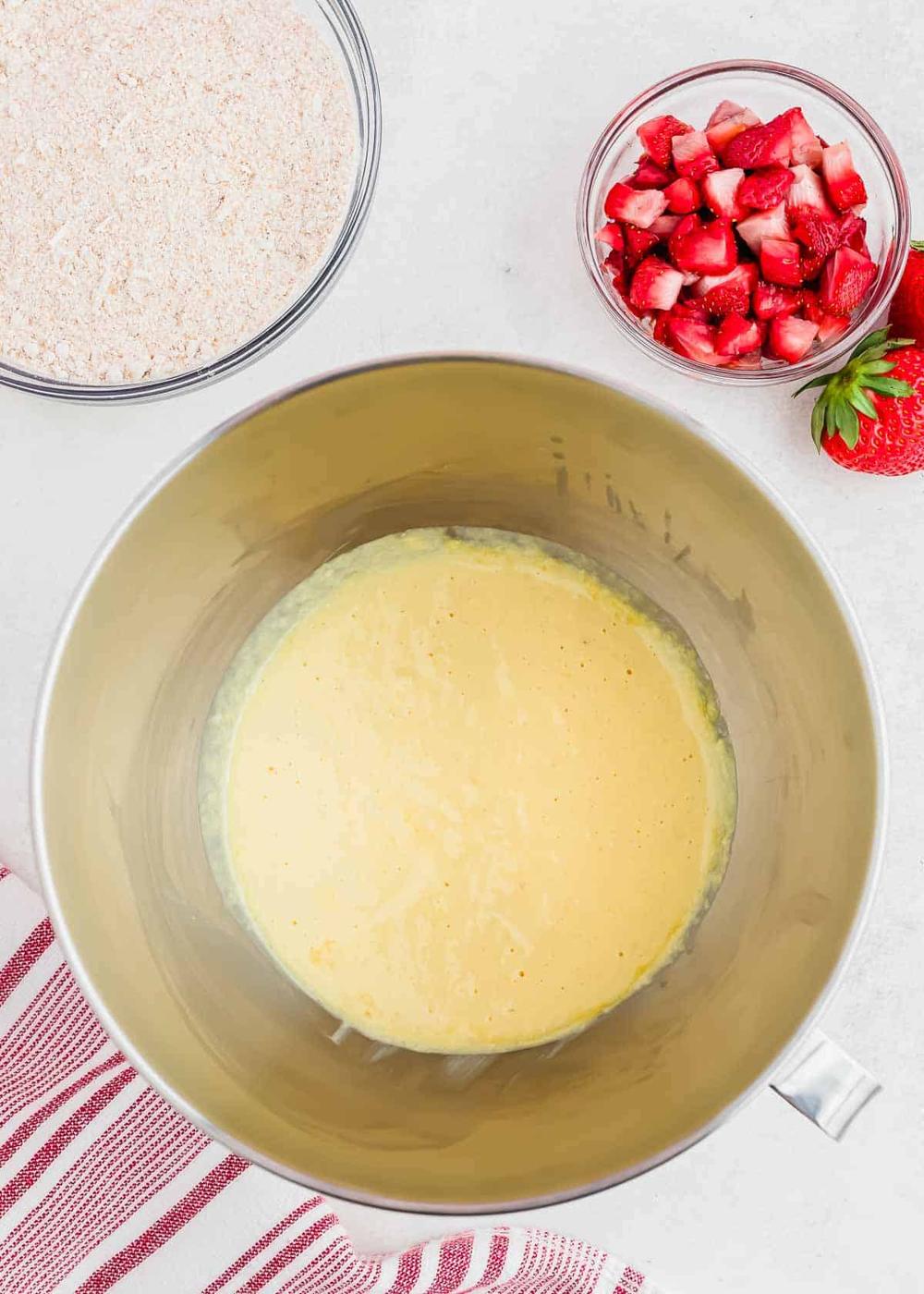 A mixing bowl with a yellow bread batter is placed on a countertop. Next to it, there are small bowls containing chopped strawberries and a dry ingredient mixture. A red and white striped cloth is partially visible in the corner.