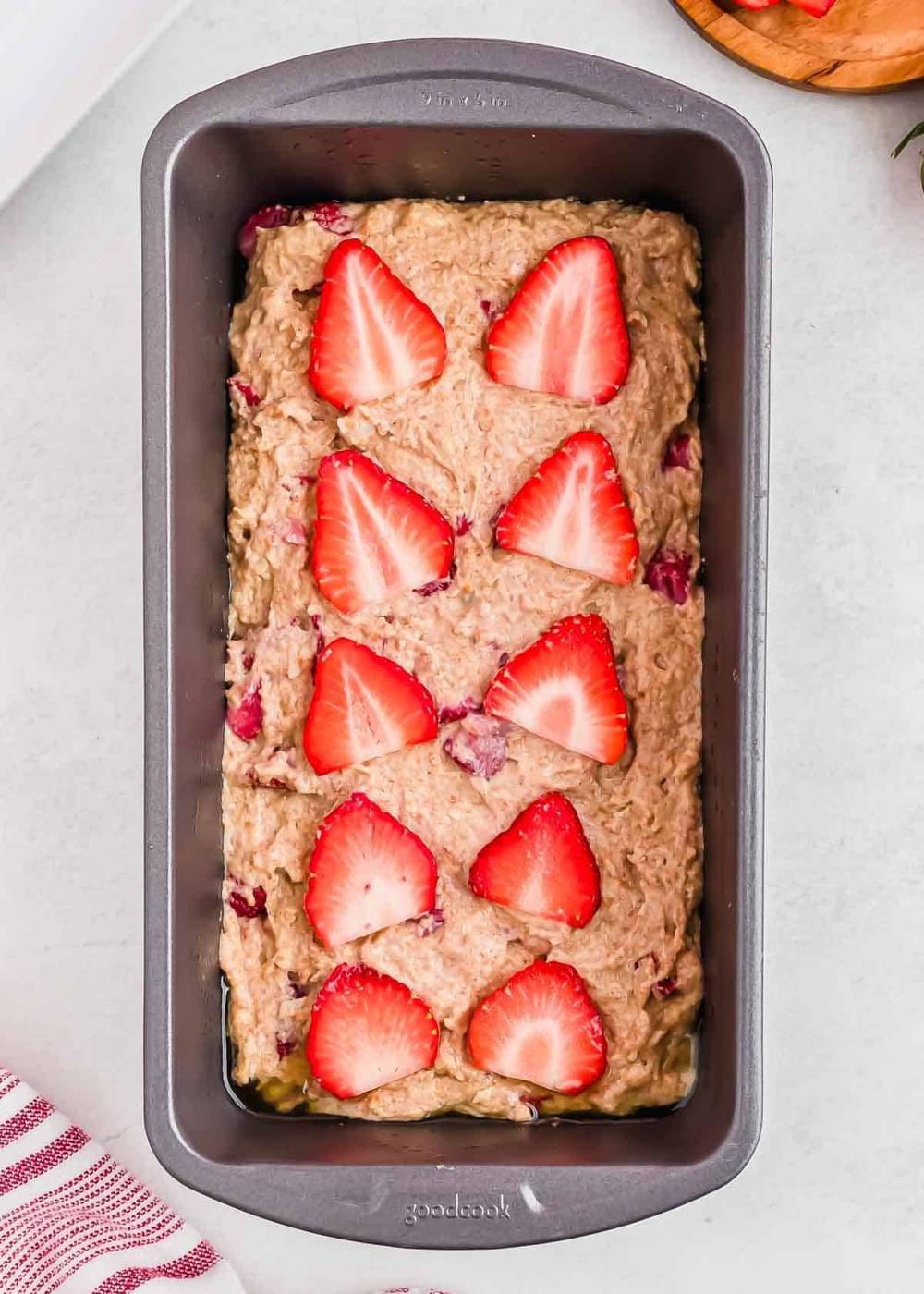 Loaf pan filled with unbaked strawberry bread batter, topped with sliced strawberries in a pattern on top. The pan is placed on a light surface, and a striped cloth is partially visible in the bottom left corner.