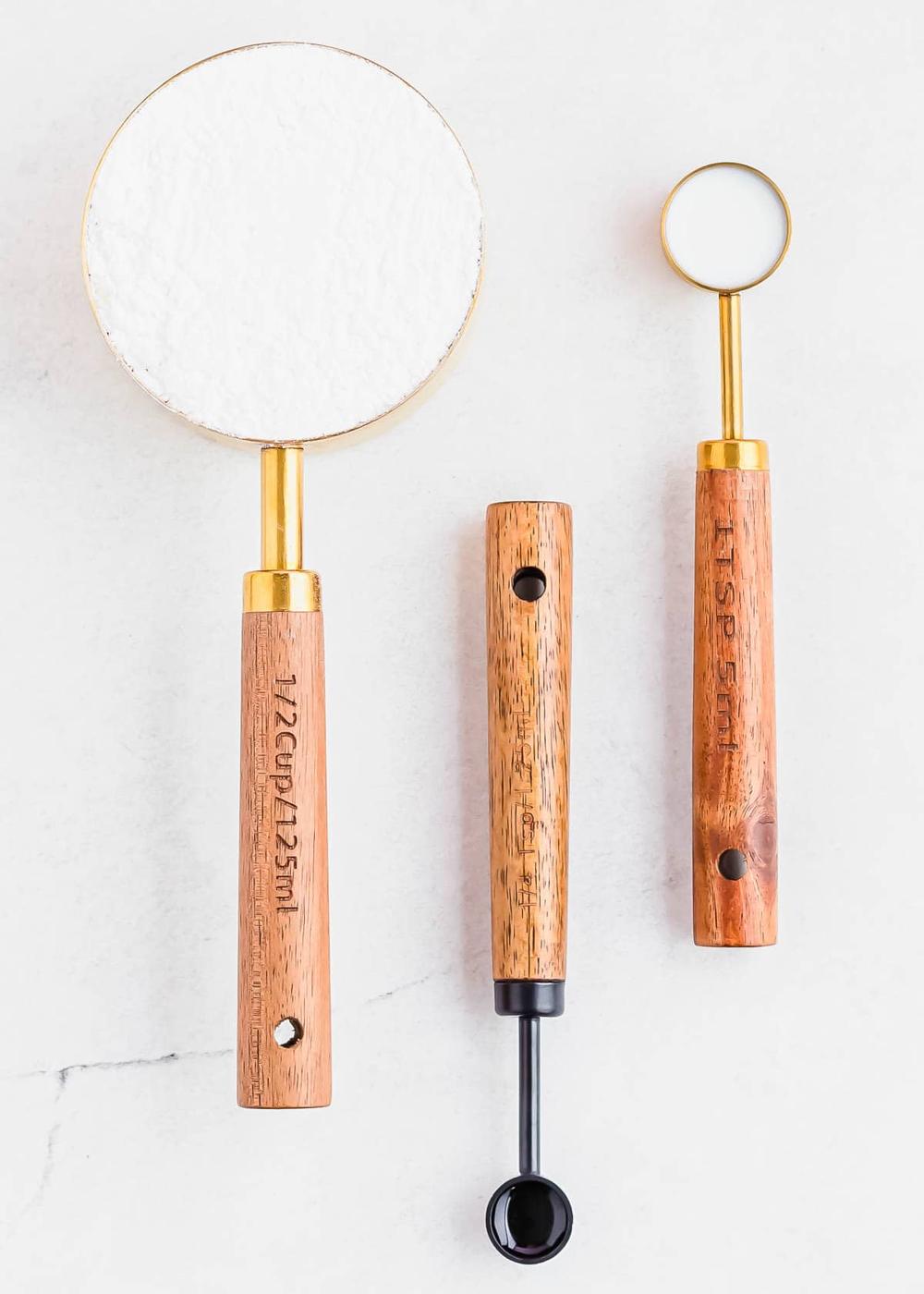 Three measuring spoons with wooden handles are arranged on a white surface. The largest contains powdered sugar, while the other two contain milk and vanilla to make a glaze. Each spoon handle has a hole for hanging and measurement markings etched into the wood.