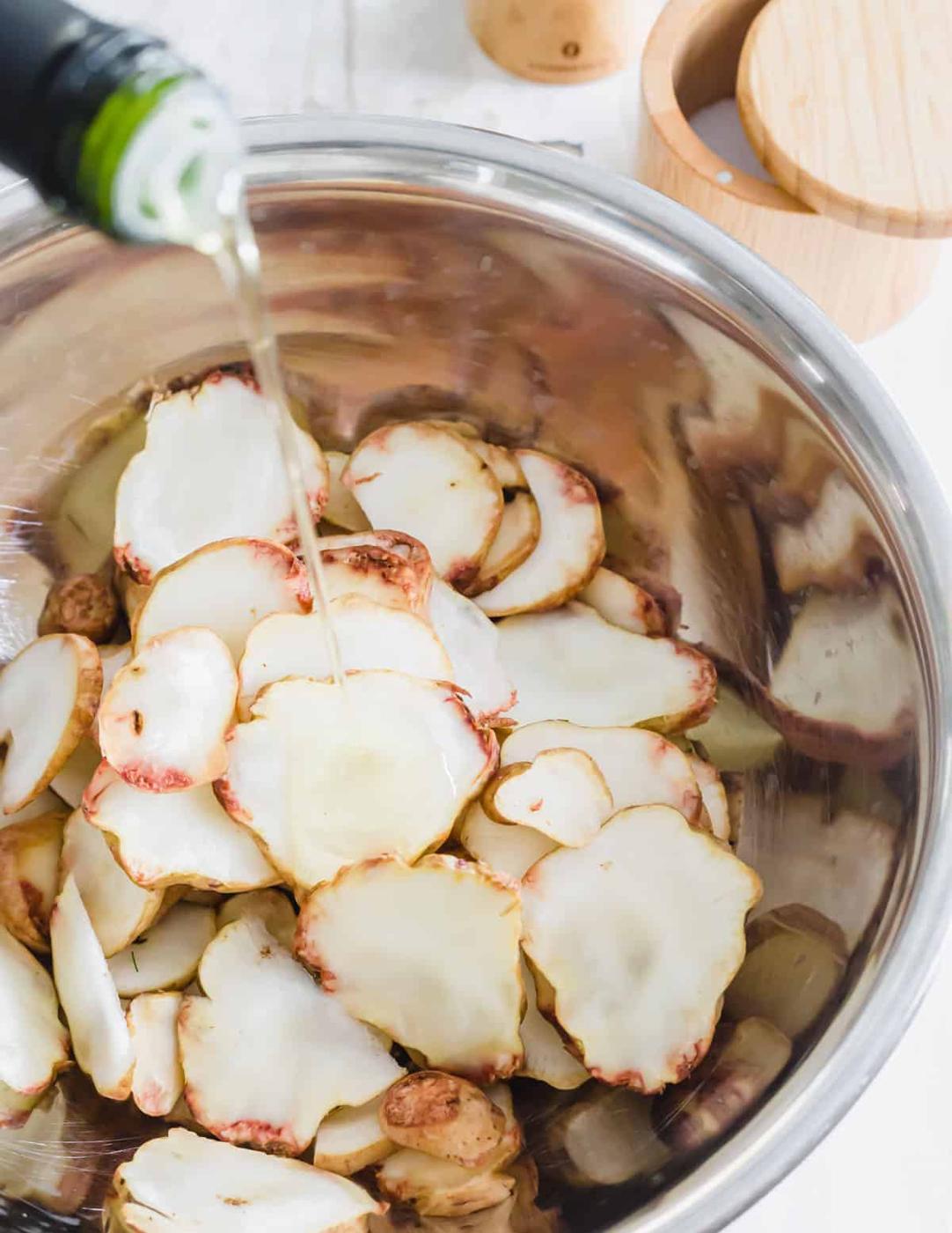 Pouring avocado oil into a large metal bowl with thinly sliced sunchokes.