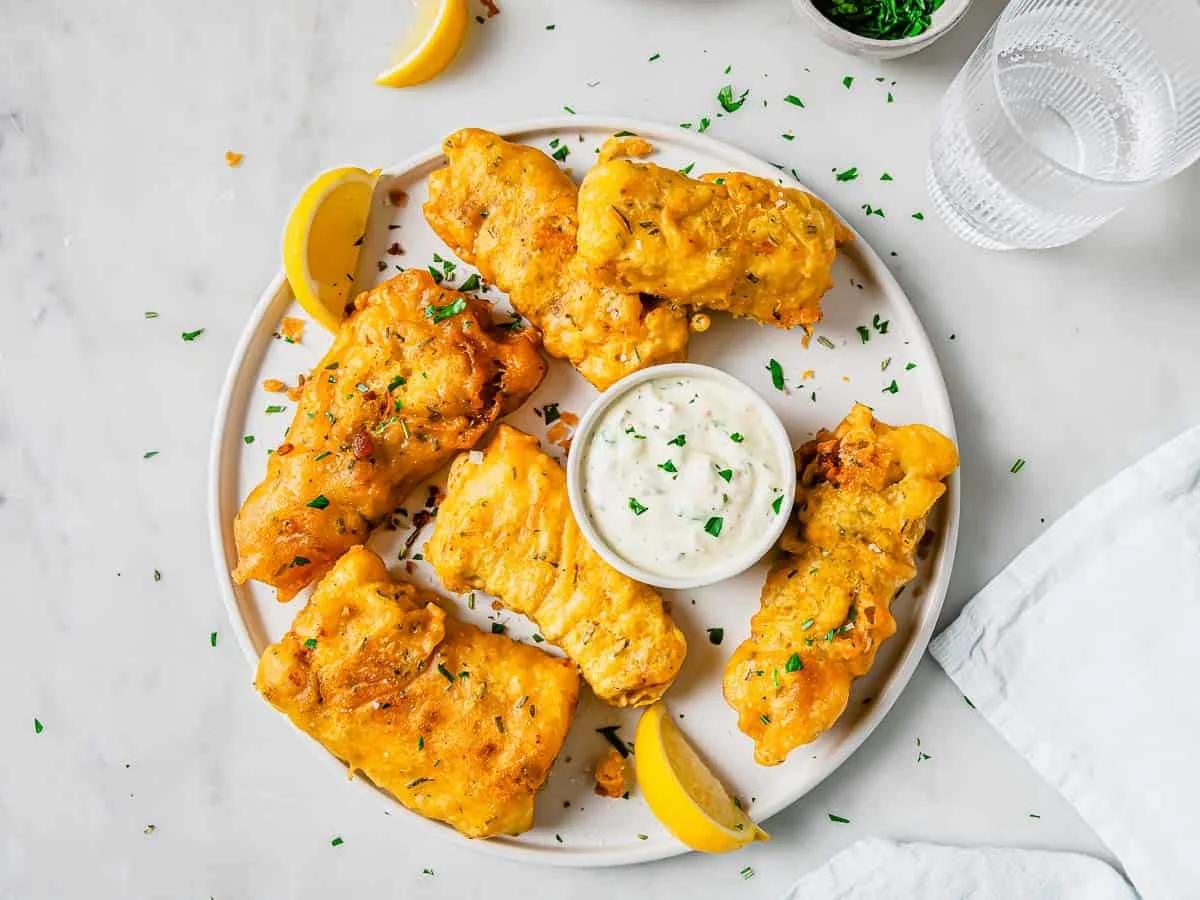 A plate of golden, crispy beer battered fried fish pieces garnished with chopped herbs and lemon wedges. A small bowl of creamy dipping sauce sits in the center. A glass of water and napkin are nearby on a light marble surface.