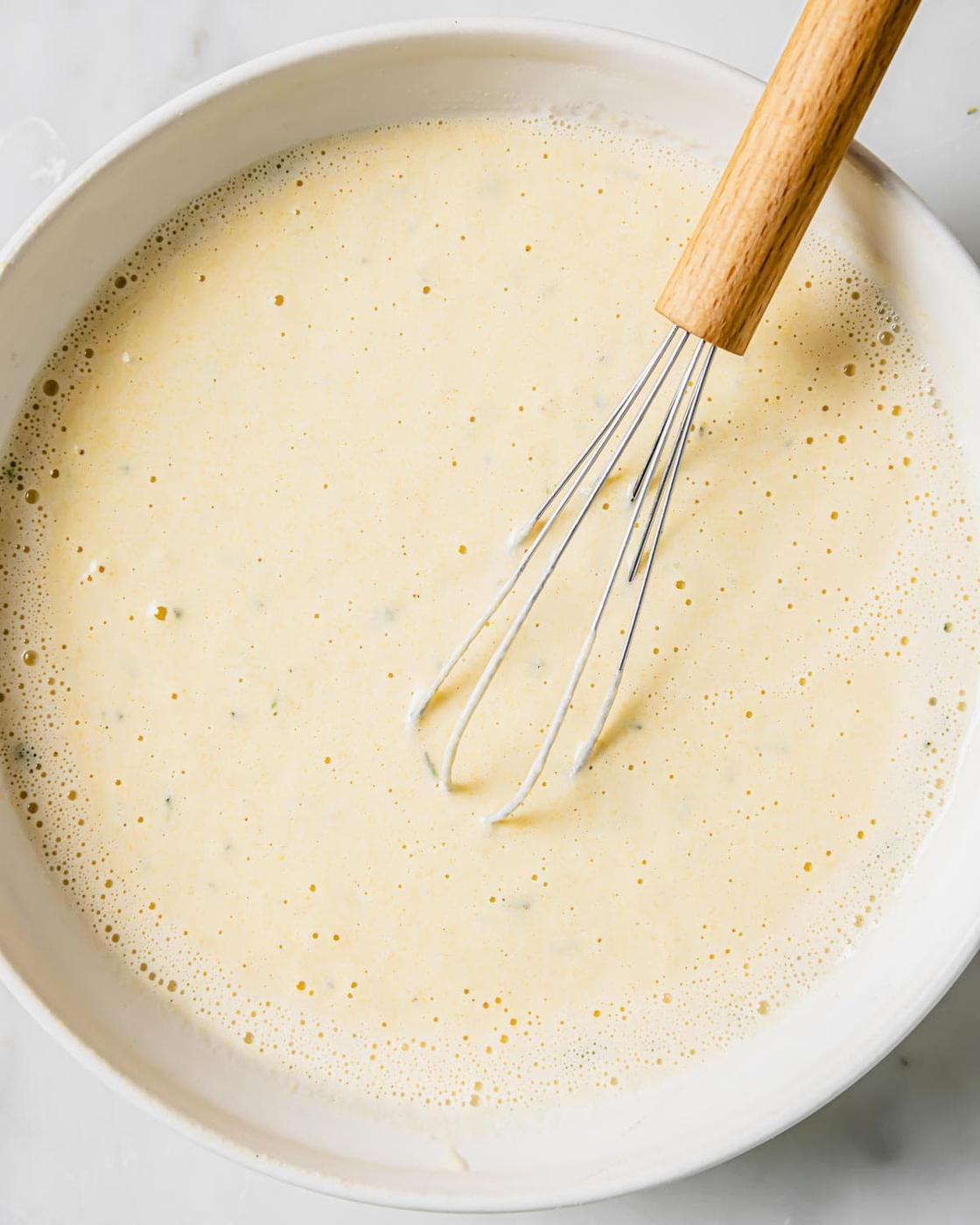 A mixing bowl filled with creamy beer batter, with air bubbles visible on the surface. A metal whisk with a wooden handle rests inside the bowl. The background is a smooth, light-colored surface.