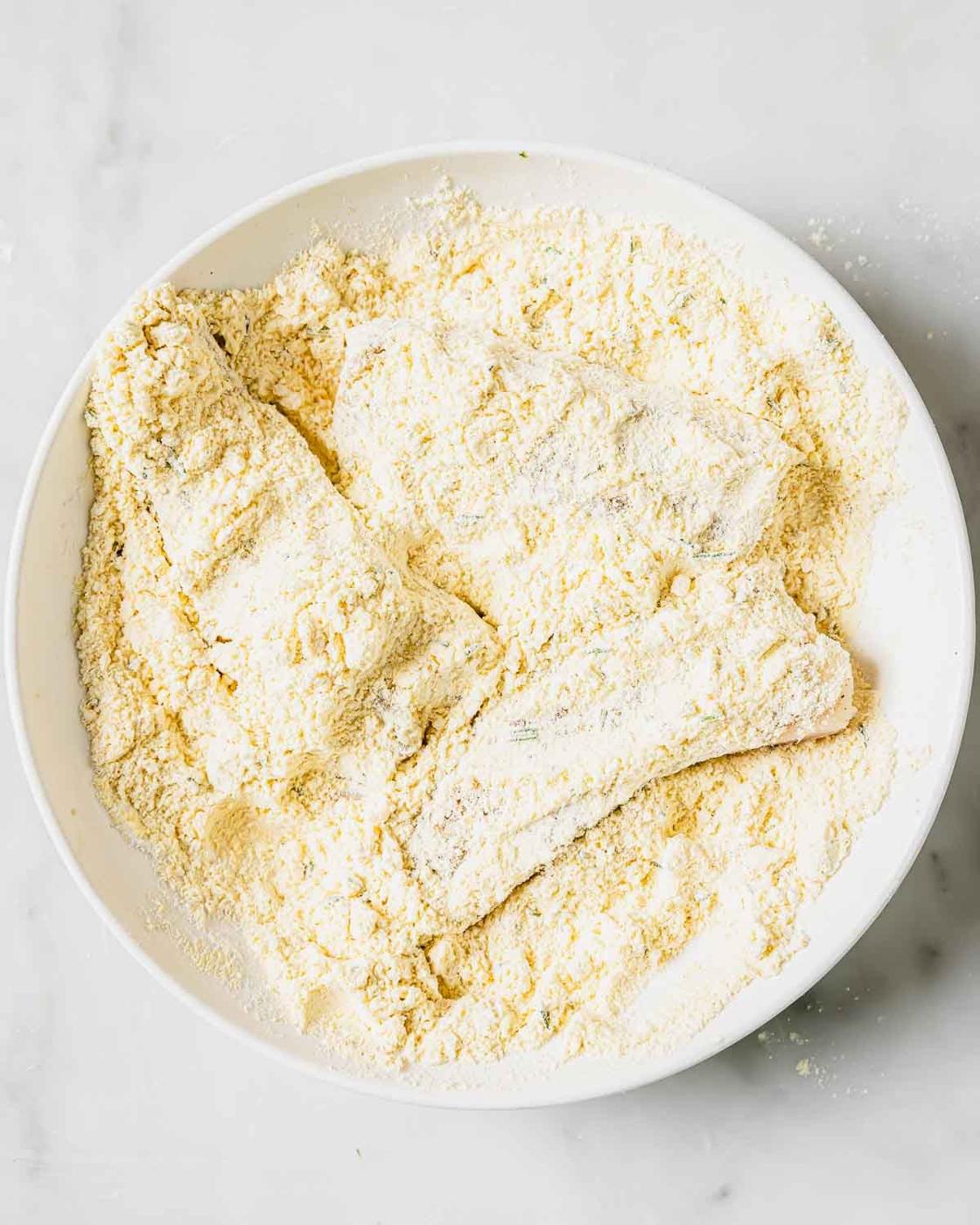 A white bowl on a marble surface contains raw fish fillets coated in a thick layer of white flour and cornmeal mixture, ready for frying.