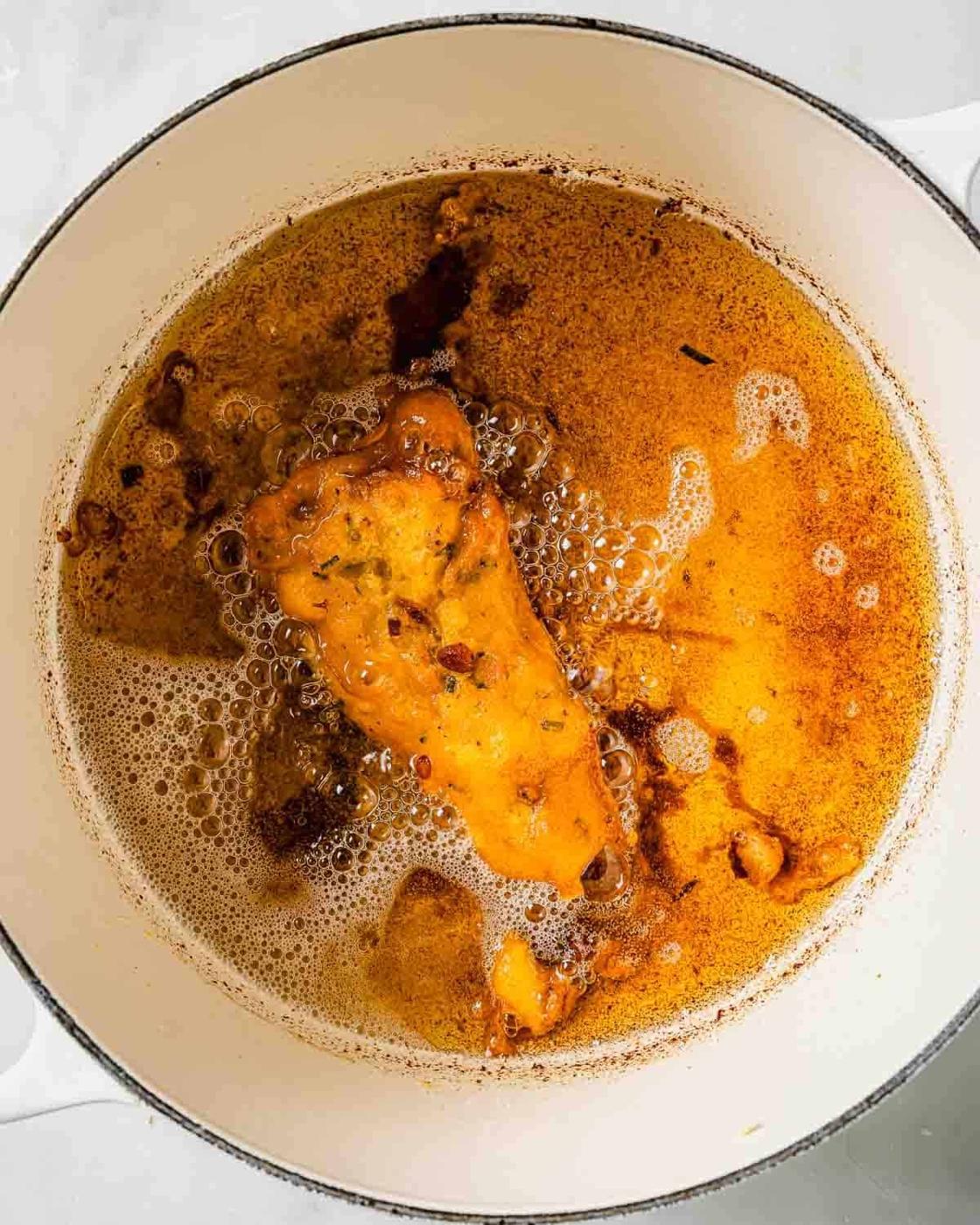 A piece of fried fish cooking in hot, bubbling oil inside a white pot. The fish is golden and crispy, surrounded by small burnt bits and foam in the oil, indicating it is in the middle of frying.