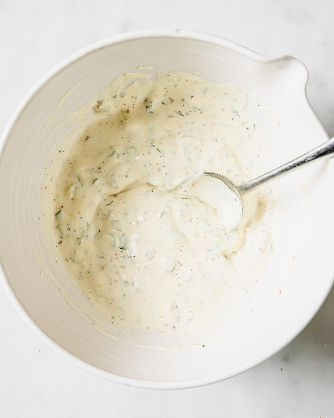 A white mixing bowl filled with a creamy, remoulade sauce. A spoon rests inside, suggesting stirring. The mixture appears to have herbs and spices blended in, visible as small green and brown flecks. The background is a smooth white surface.