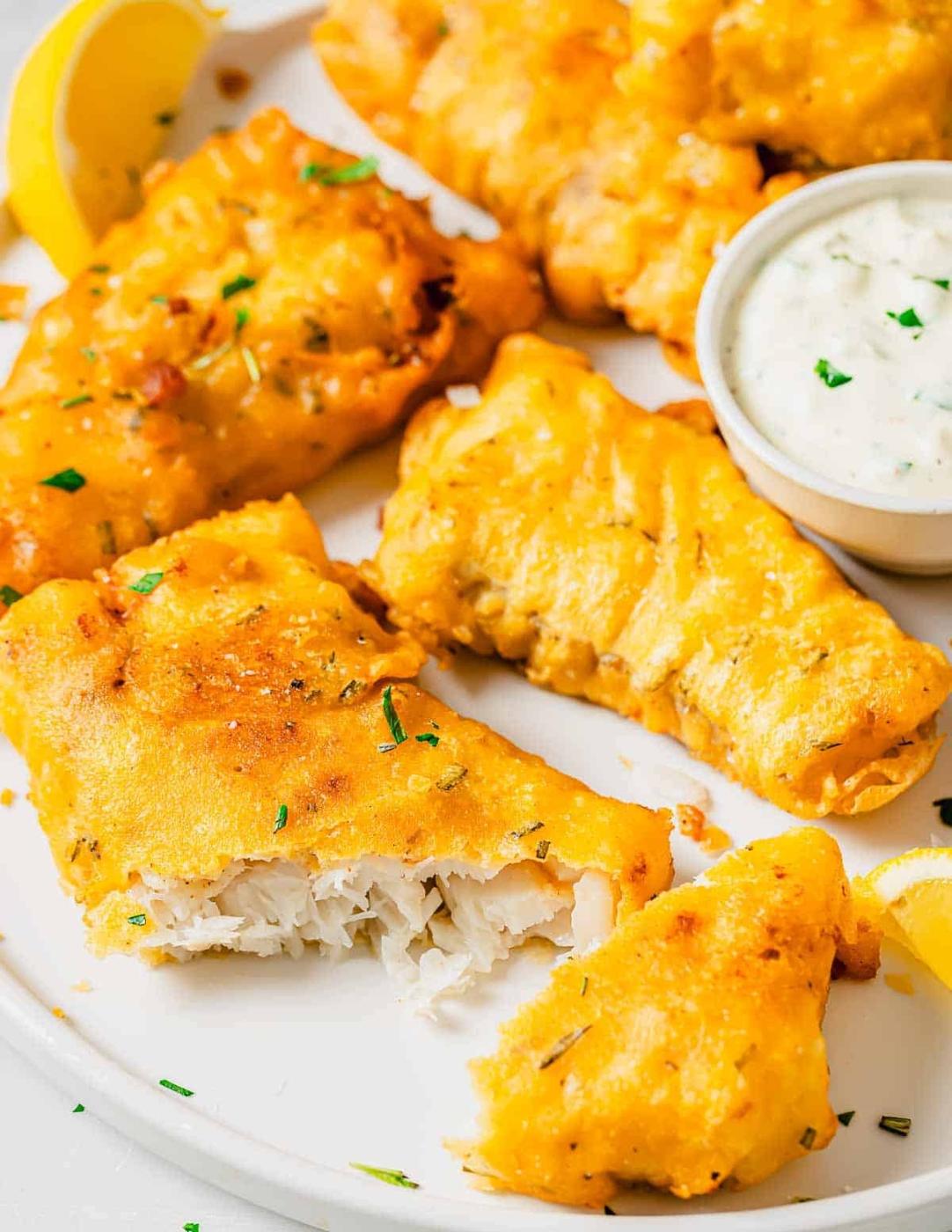 Golden-brown battered fish pieces on a white plate, garnished with chopped herbs. Some pieces show flaky white fish inside. A small bowl of creamy dipping sauce and lemon wedges are beside the fish.
