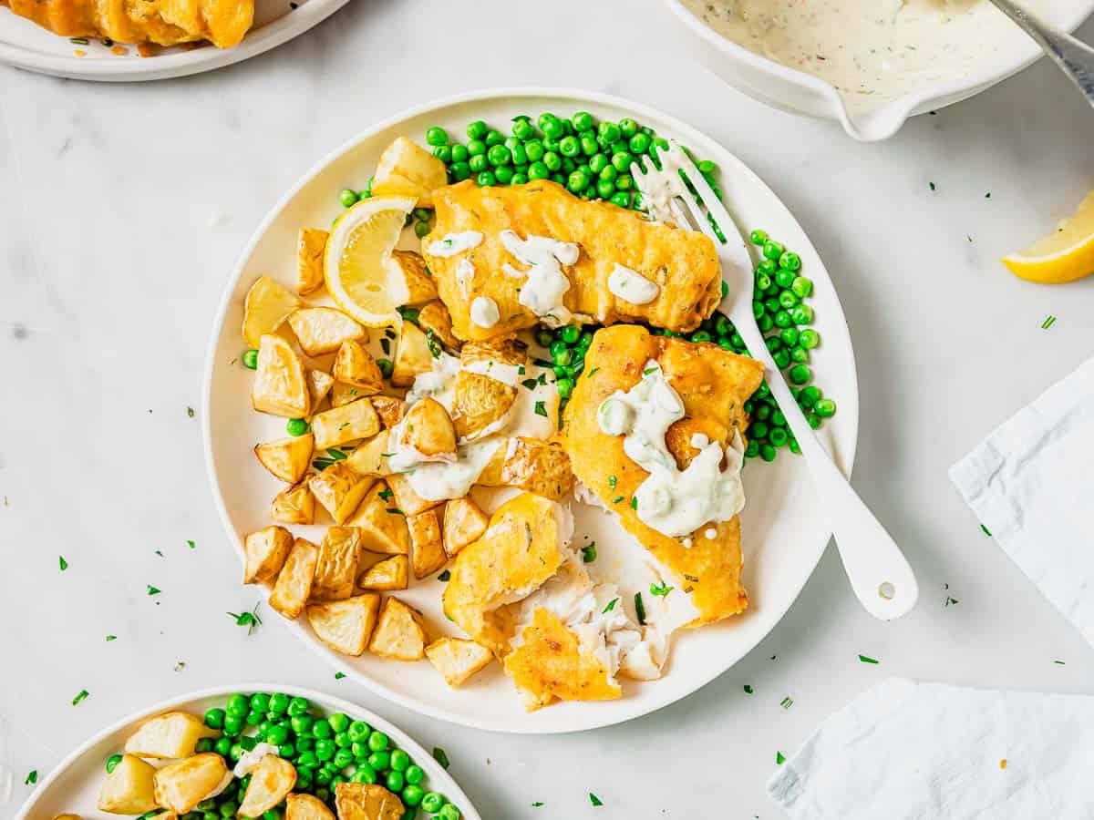A plate of crispy golden beer battered fish fillets with tartar sauce, served with roasted potatoes, bright green peas, and lemon wedges. A fork rests on the plate, and a bowl of extra sauce is nearby on a white background.