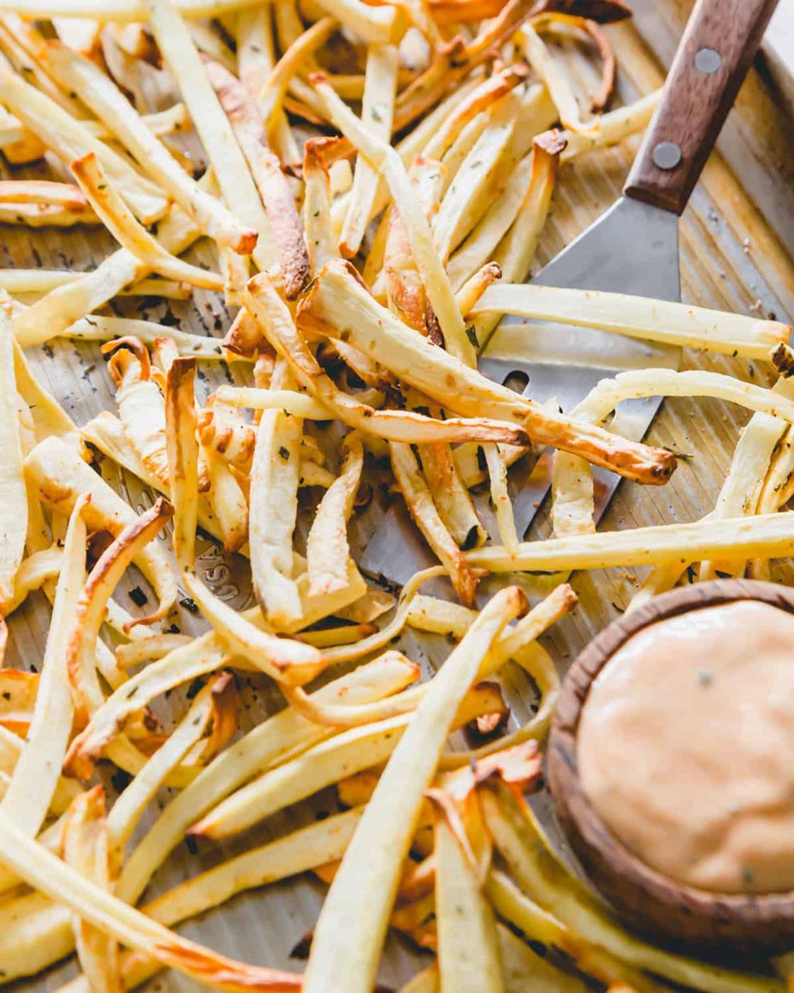 Crispy golden brown parsnip fries straight out of the air fryer and oven on a baking sheet with fry sauce and a spatula for serving.