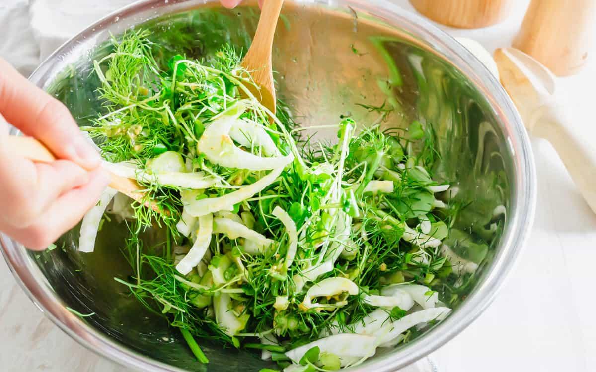 Tossing raw sliced fennel and baby arugula in a bowl with wooden salad utensils.