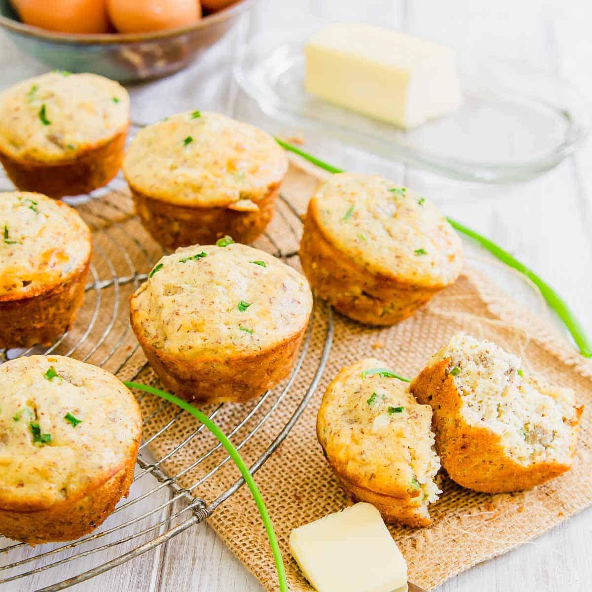 A cooling rack with six savory sausage muffins garnished with herbs. A seventh muffin is halved with a pat of butter beside it. A block of butter and a bowl of eggs are in the background.