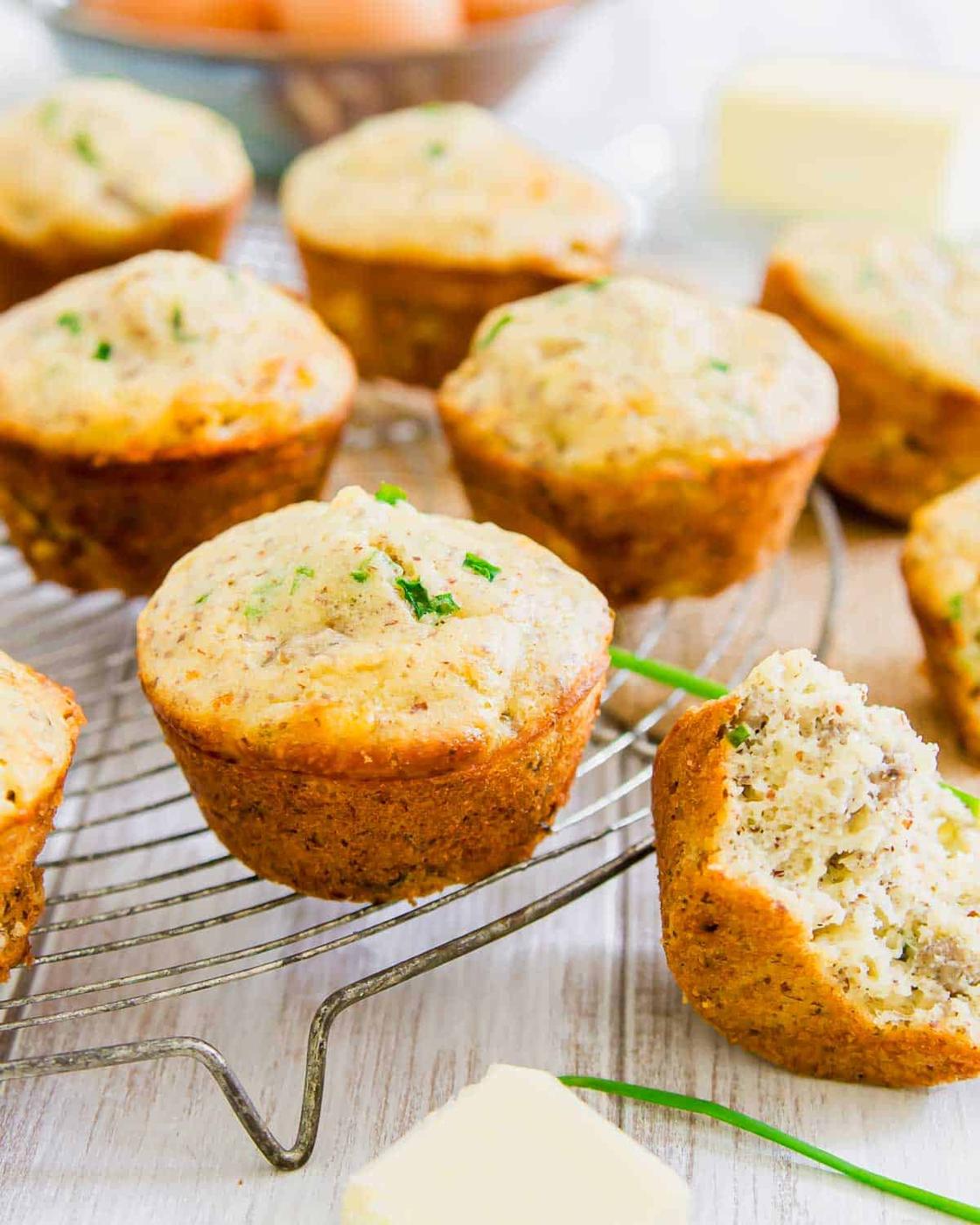 Cooling rack with freshly baked sausage muffins garnished with herbs. One muffin is partially eaten, revealing a moist interior. Butter cubes are placed on the side.