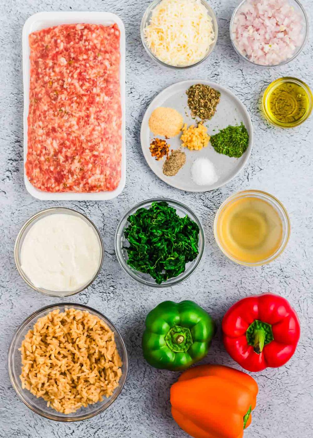 Ingredients for a sausage stuffed pepper recipe laid out on a gray surface, including ground Italian sausage, cheese, chopped onions, spinach, bell peppers, rice, various seasonings and oil.