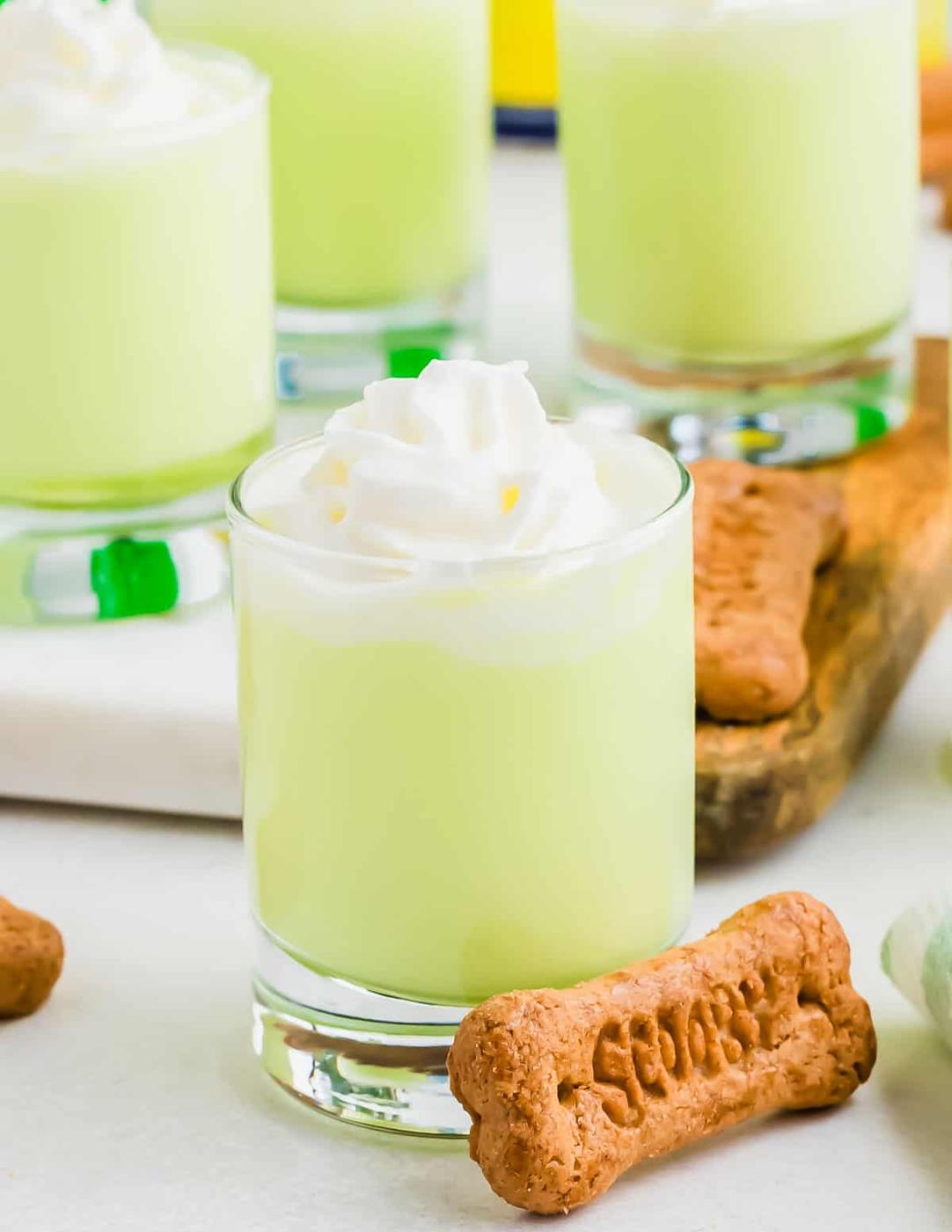 Four glasses of Scooby shot drinks topped with whipped cream are arranged next to dog bone-shaped biscuits that say Scooby on a white surface.
