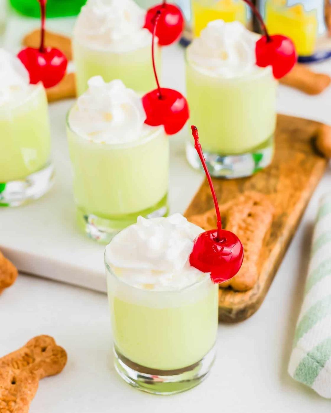 Shot glasses filled with Scooby snack shot drink recipe with whipped cream and a cherry are arranged on a table, alongside dog-bone shaped graham cracker cookies and a striped cloth.