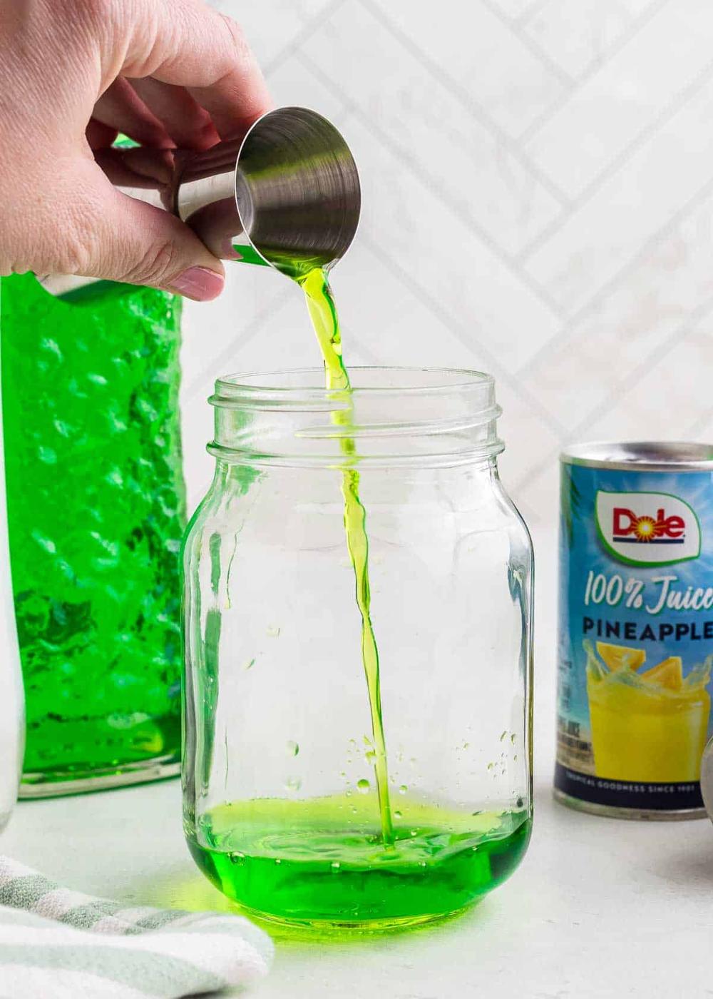 Hand pouring Midori melon from a jigger into a clear mason jar, with a bottle of green liquid and a can of pineapple juice visible in the background.