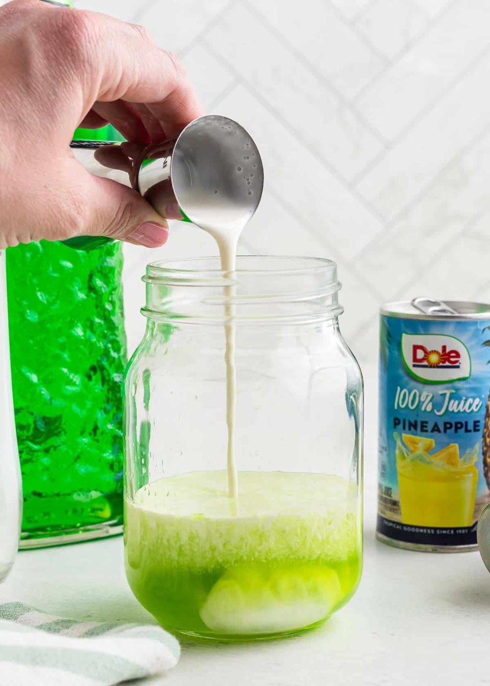 A hand pours cream from a measuring cup into a glass jar containing a green liquid. A green bottle and a can of pineapple juice are in the background.