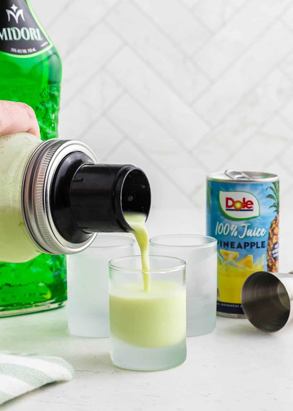 A person pouring a Scooby snack shot drink from a cocktail shaker into a shot glass. A green bottle and a can of Dole pineapple juice are on the counter beside two frosted glasses.