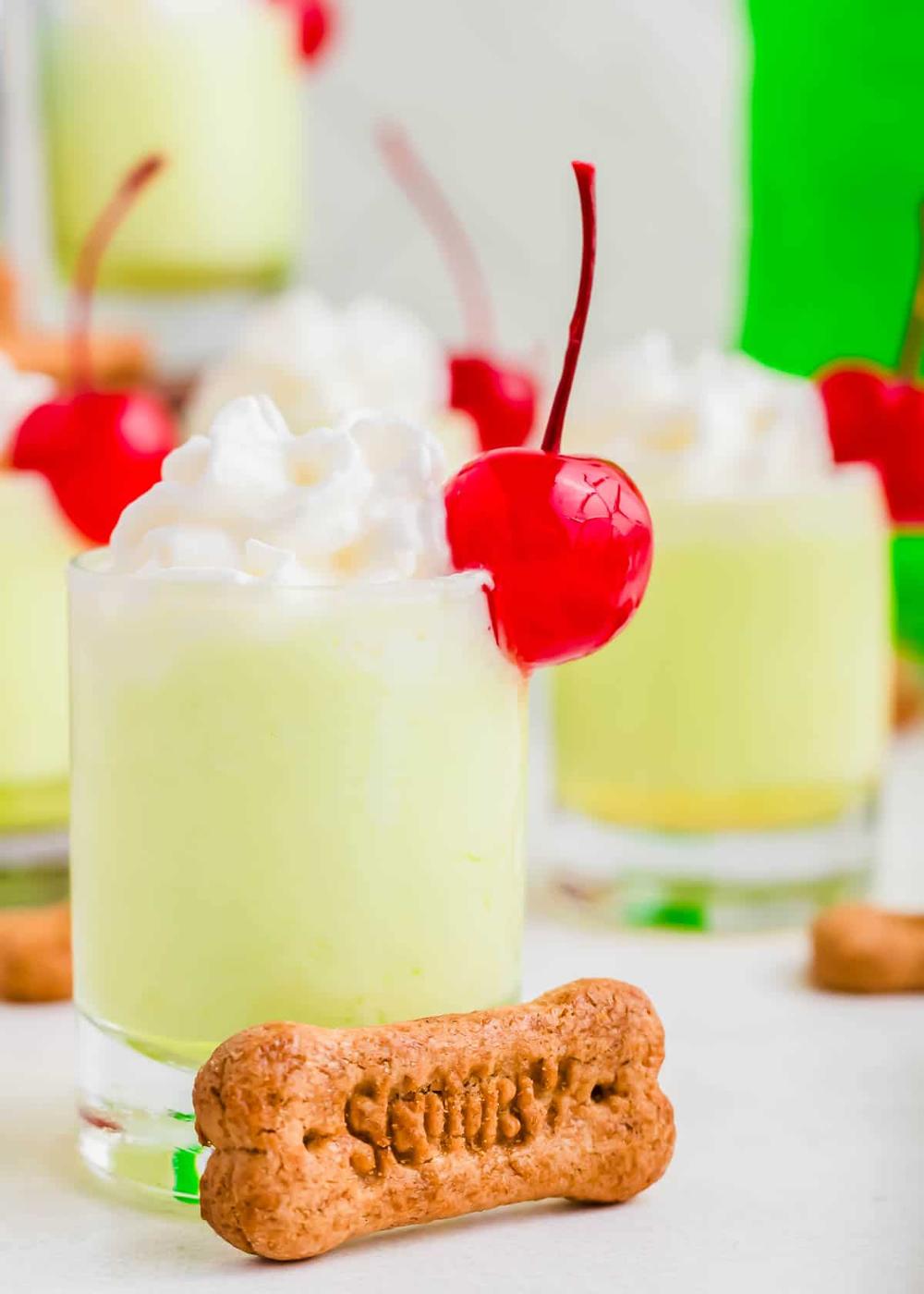 A creamy green drink topped with whipped cream and a cherry in a small glass, accompanied by a bone-shaped cookie labeled 'Scooby.' Other similar drinks are in the background.