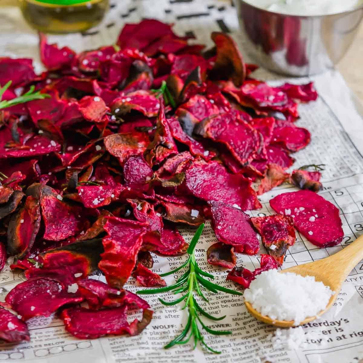 A pile of crispy red beet chips is spread on newspaper. A wooden spoon holds coarse salt beside fresh rosemary sprigs. A metal bowl of dipping sauce is partially visible in the background.