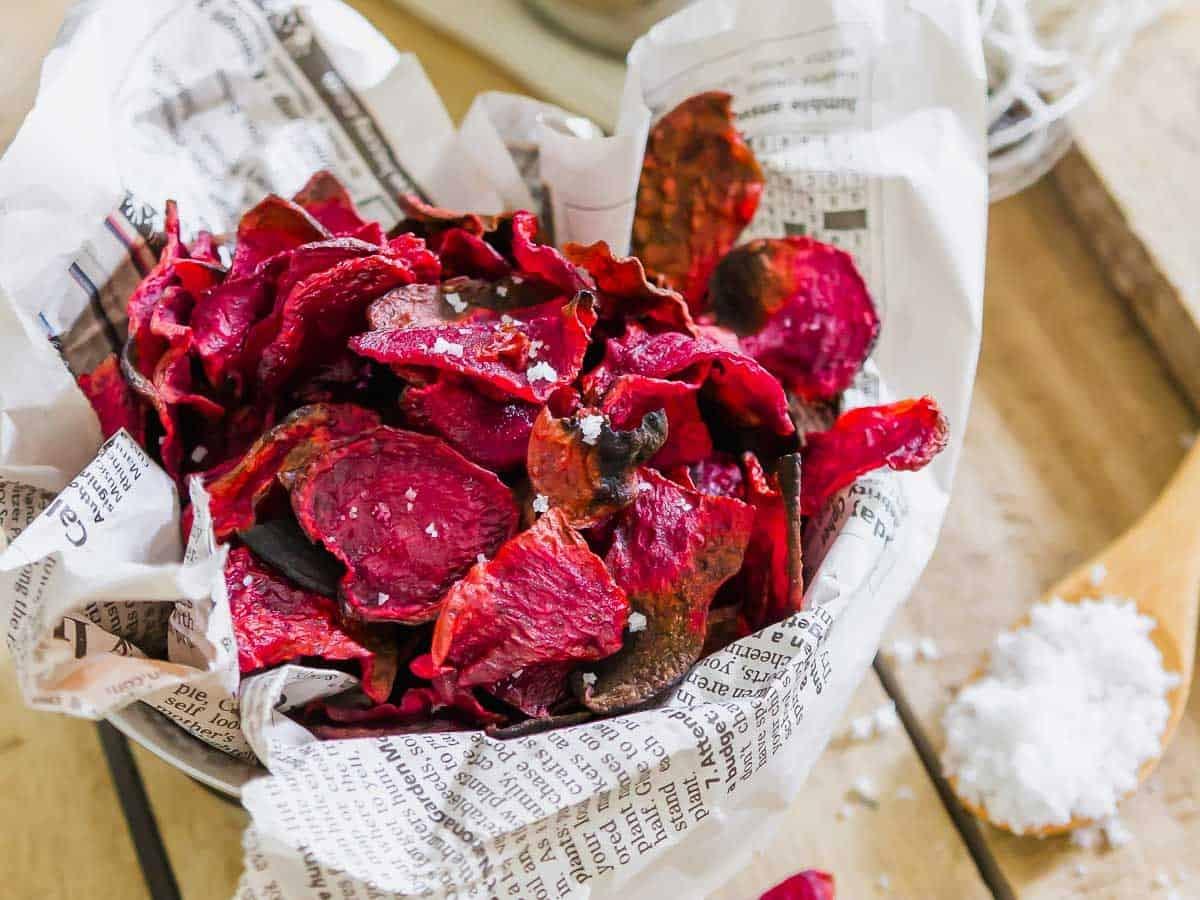 A bowl of crispy, red beet chips sits on a wooden surface. The chips are arranged in newspaper and topped with coarse sea salt, with a small pile of salt nearby. The background includes rustic kitchen elements.
