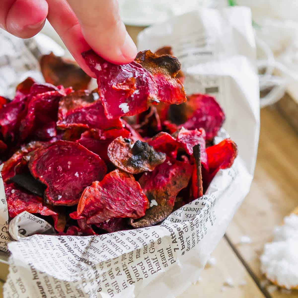 A hand is reaching for a beet chip from a bowl lined with newspaper. The vibrant red chips are sprinkled with coarse salt, and the scene is set on a wooden surface.