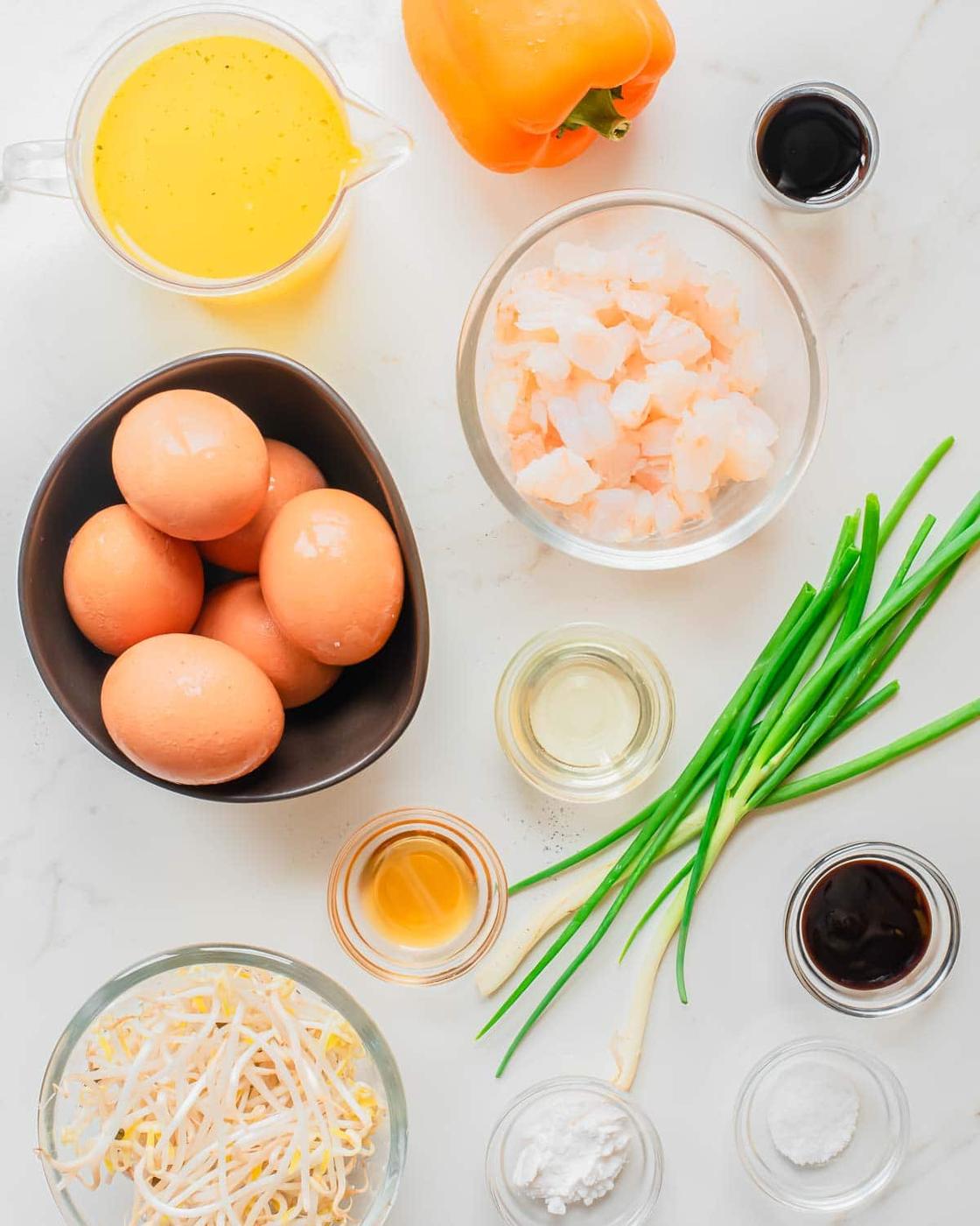 A top-down view of cooking ingredients on a white surface, including eggs, shrimp, yellow bell pepper, green onions, bean sprouts, broth, and various sauces in small bowls.