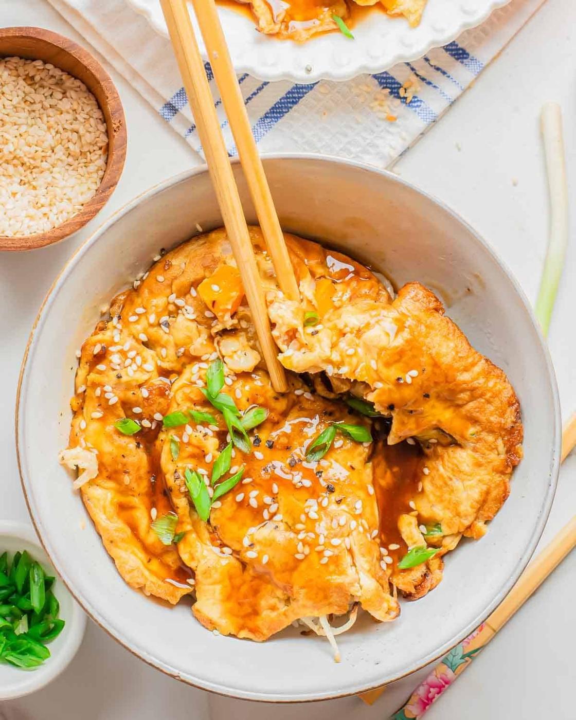 A bowl of savory Chinese omelettes covered in brown sauce, topped with sesame seeds and chopped green onions. Chopsticks are picking up a piece, and small bowls of sesame seeds and green onions are nearby.