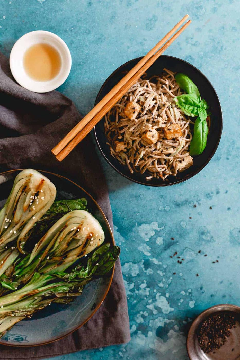 Grilled baby bok choy with sesame seeds on a dark plate with noodles and tofu next to it in a black bowl.