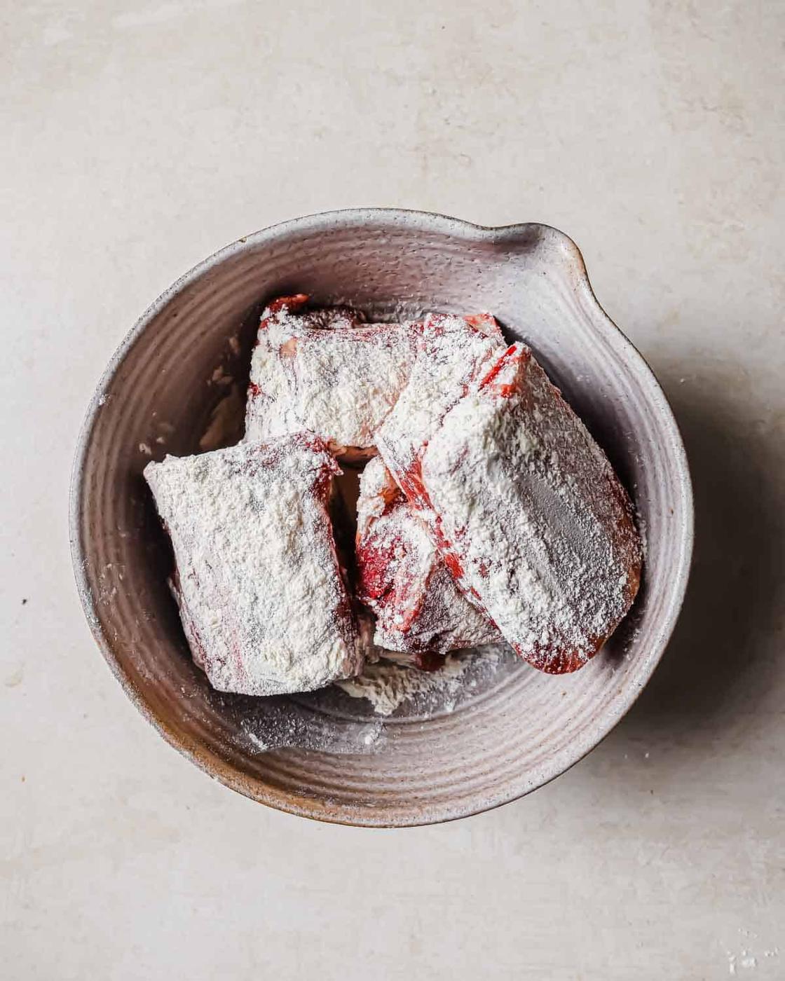 A bowl containing short rib pieces coated in flour, placed on a light-colored surface. The bowl has a rustic, textured appearance.