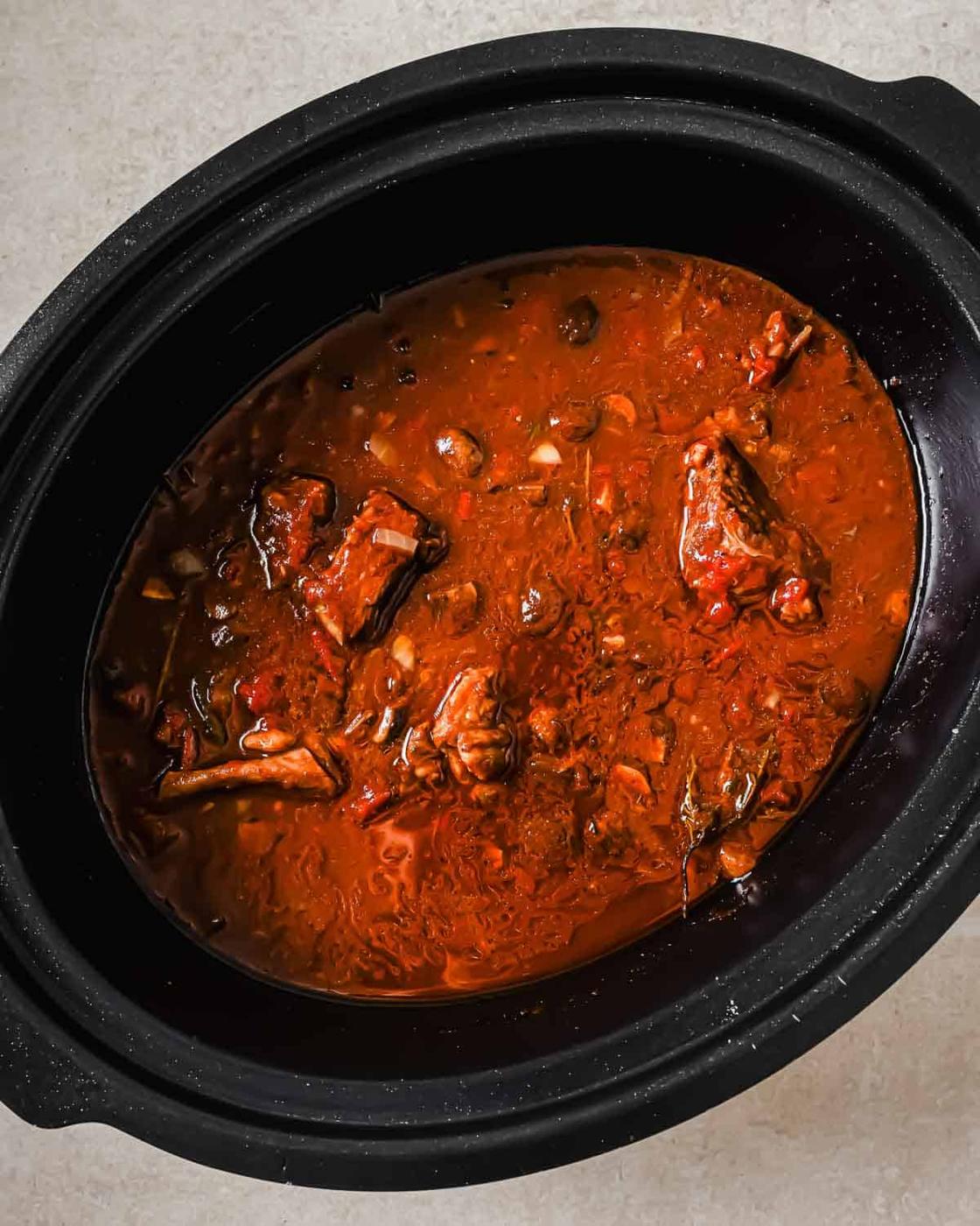 A black slow cooker filled with rich, cooked short ribs containing chunks of meat and visible small onions or garlic cloves. The stew has a thick, hearty consistency.