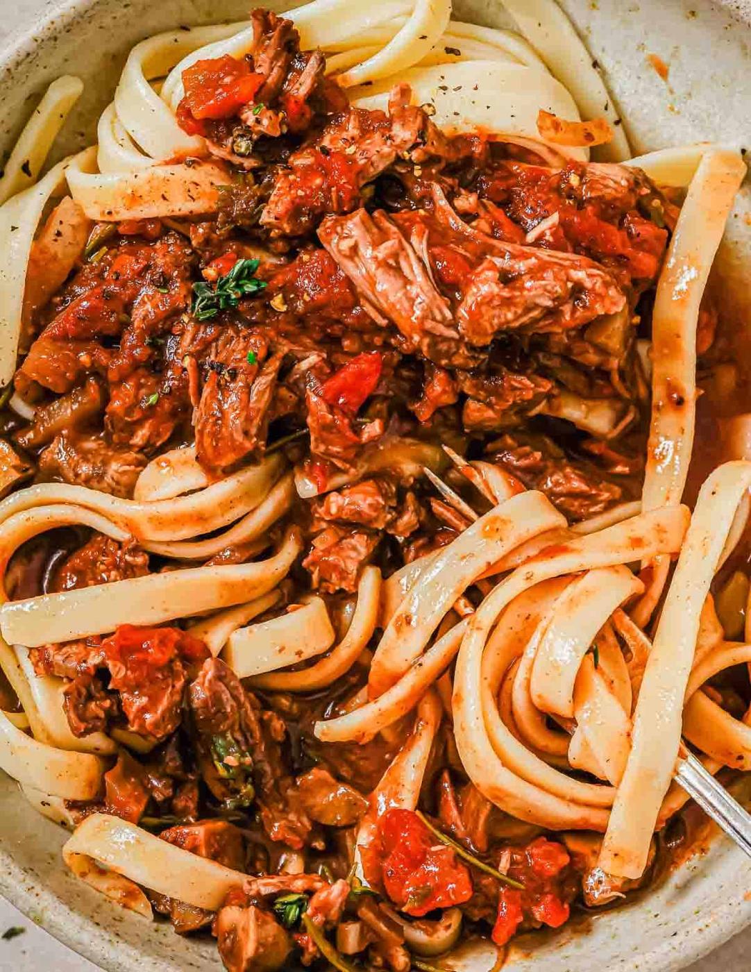 A close-up of a bowl of pasta with tender shredded short ribs and a rich, chunky tomato ragu, garnished with fresh herbs. The pasta is intertwined with the savory sauce, creating a hearty and appetizing dish.