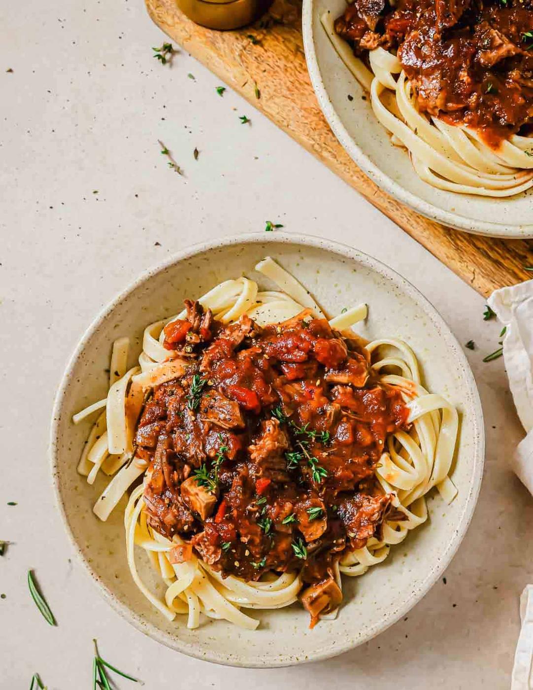 A bowl of pasta topped with short rib ragu, garnished with fresh herbs. The dish is set on a light-colored surface, with another bowl partially visible in the background.